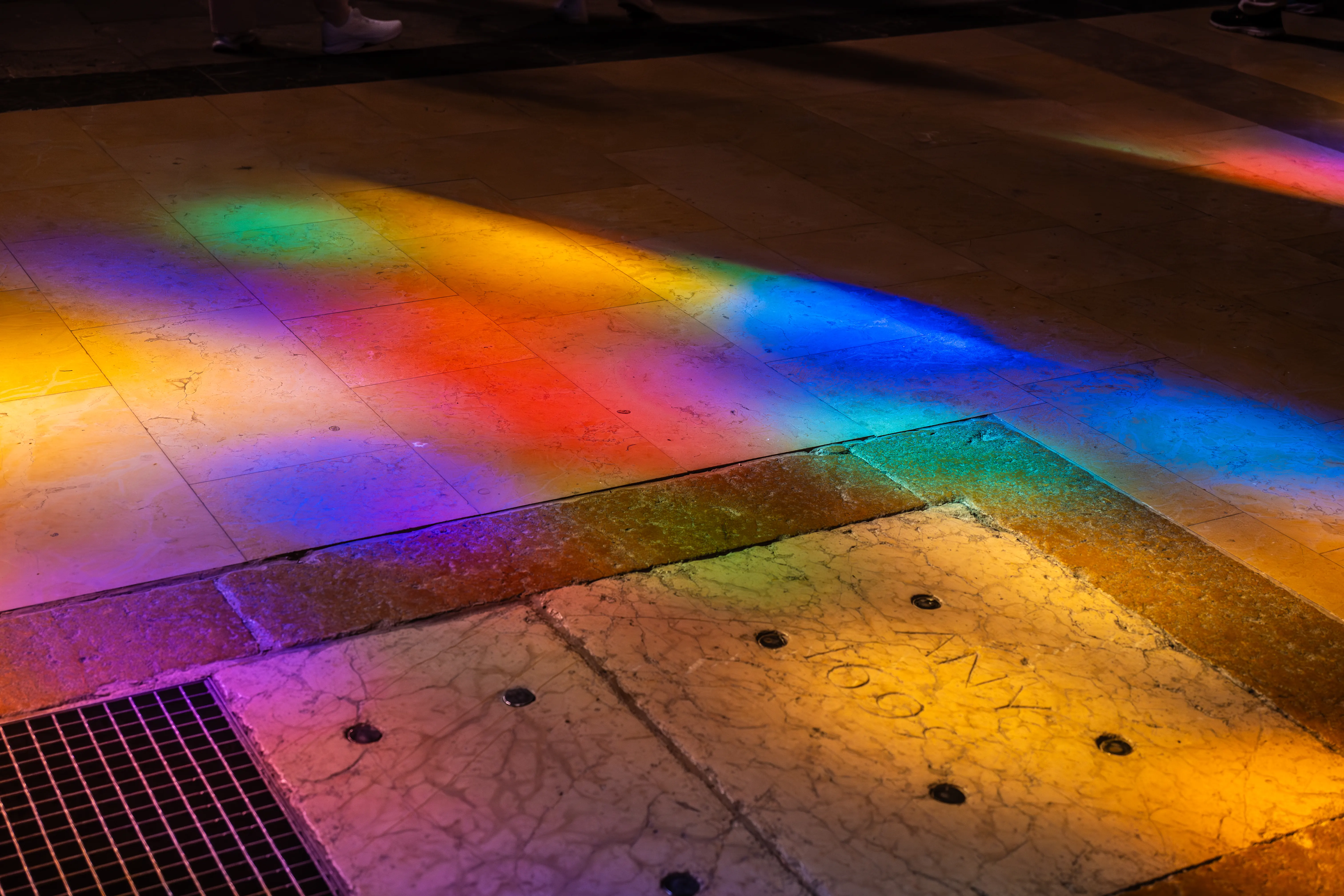 A floor made of stone and tile is illuminated by colourful light beams in shades of yellow, orange, red, purple, blue, and green. A metal grate is visible in the lower left corner.
