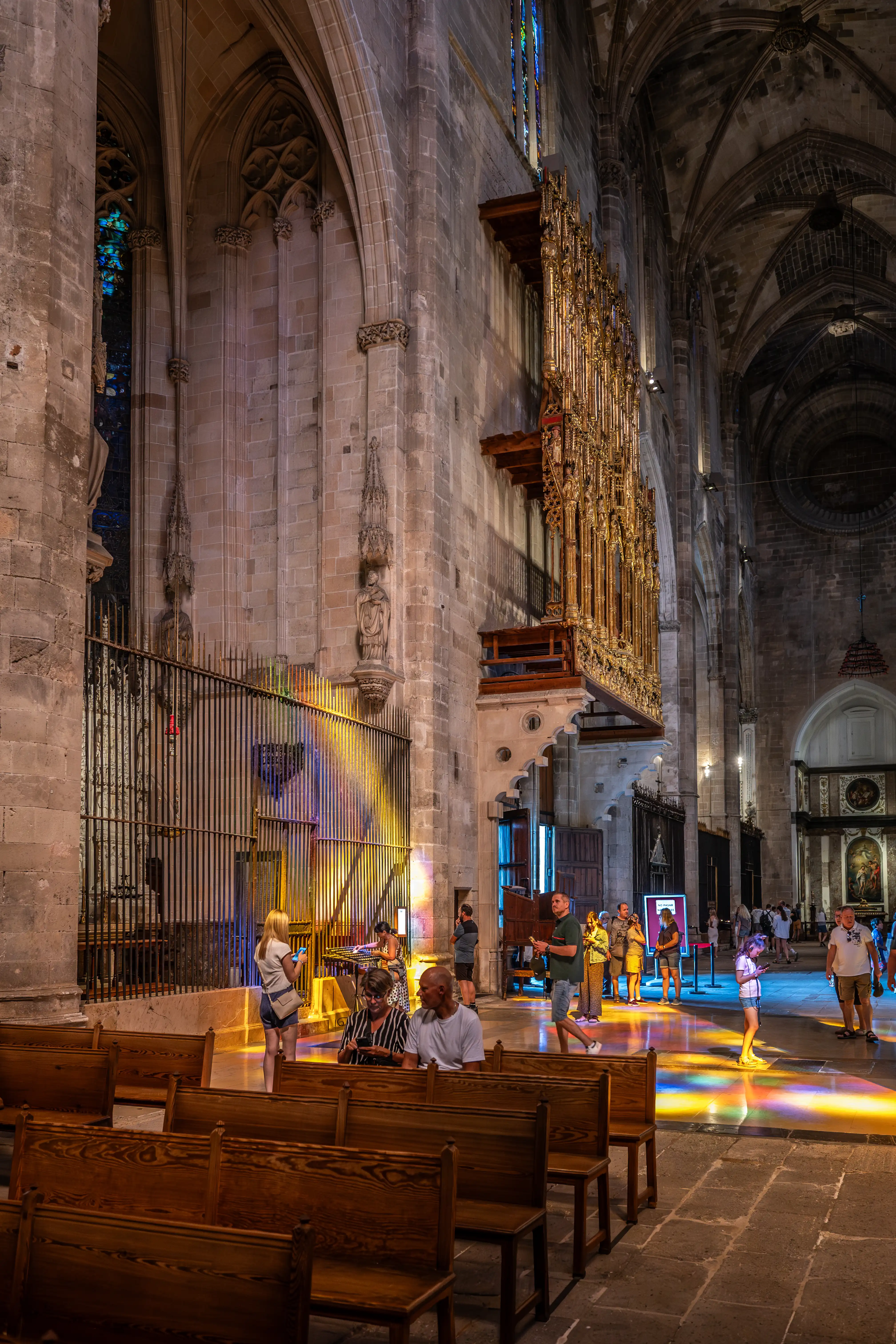 Interior of a cathedral showing a large, golden pipe organ mounted on a stone wall. Sunlight creates a spectrum of colours on the floor and a metal barrier in front of a stained glass window. Several visitors are in the scene, with wooden pews in the foreground.