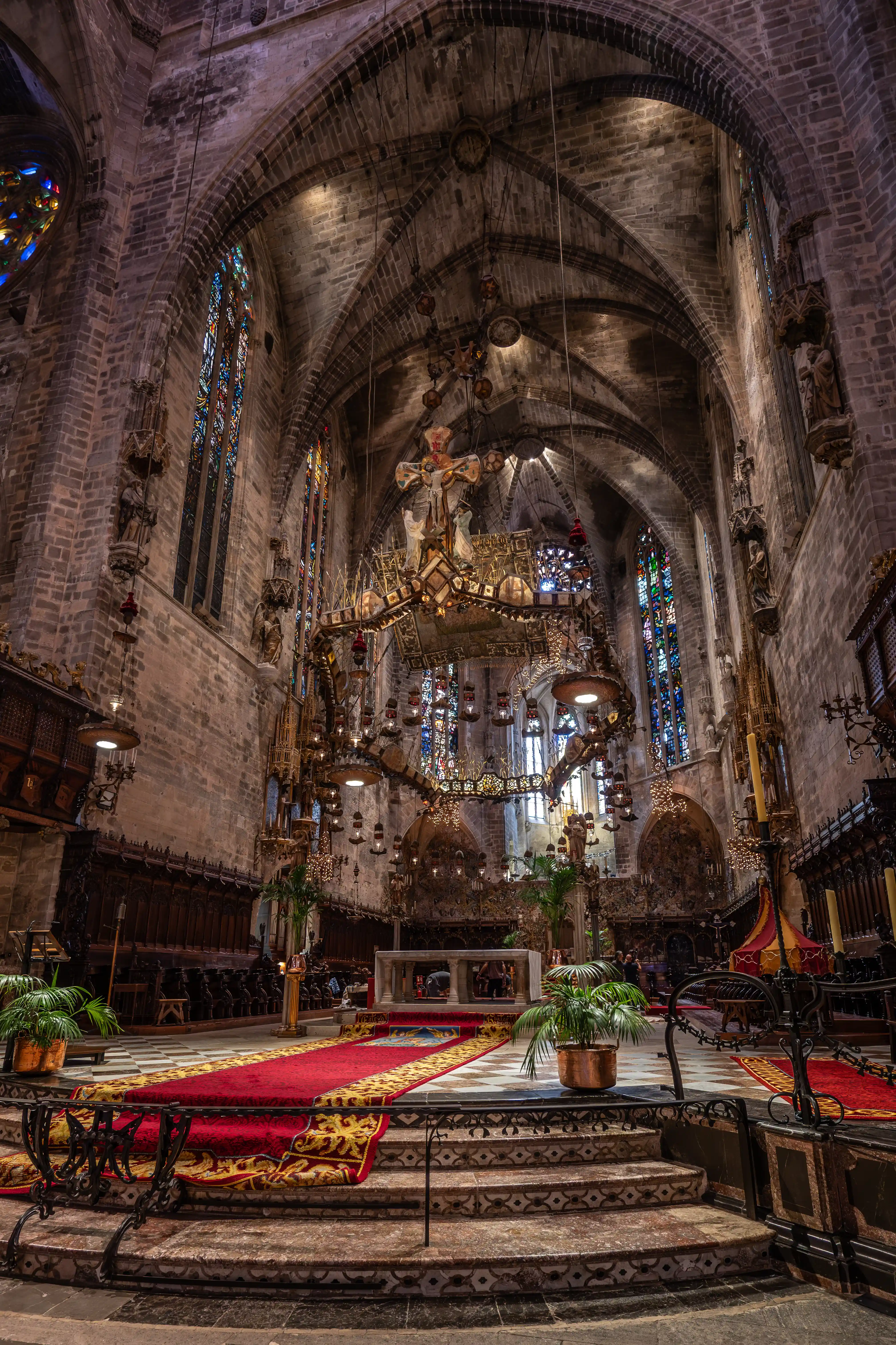 Interior of a Gothic cathedral featuring a large ornate chandelier, colourful stained glass windows, and a red carpet leading to the altar.