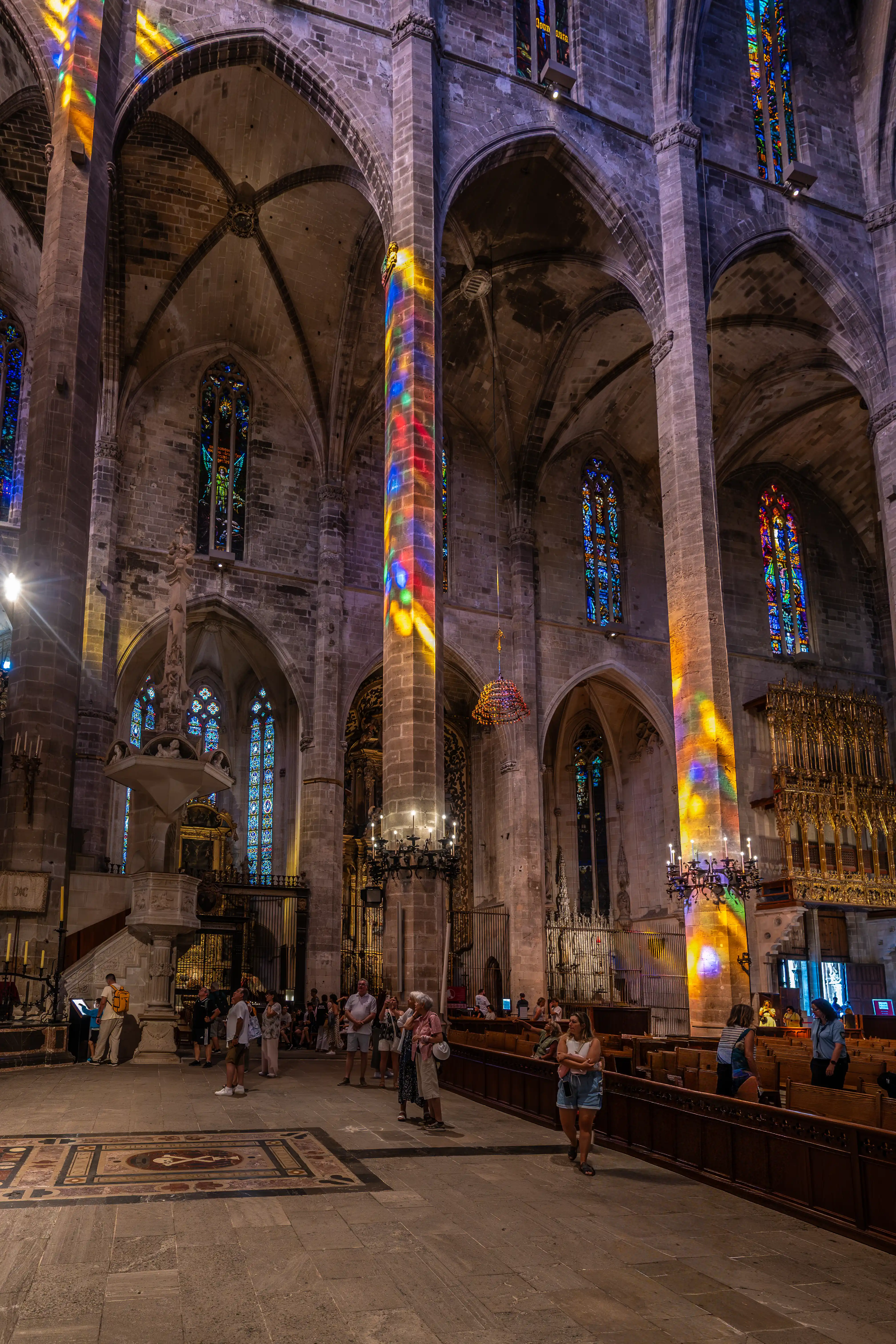 Interior of a Gothic cathedral with columns illuminated by colourful light from stained-glass windows, with tourists walking on the stone floor.