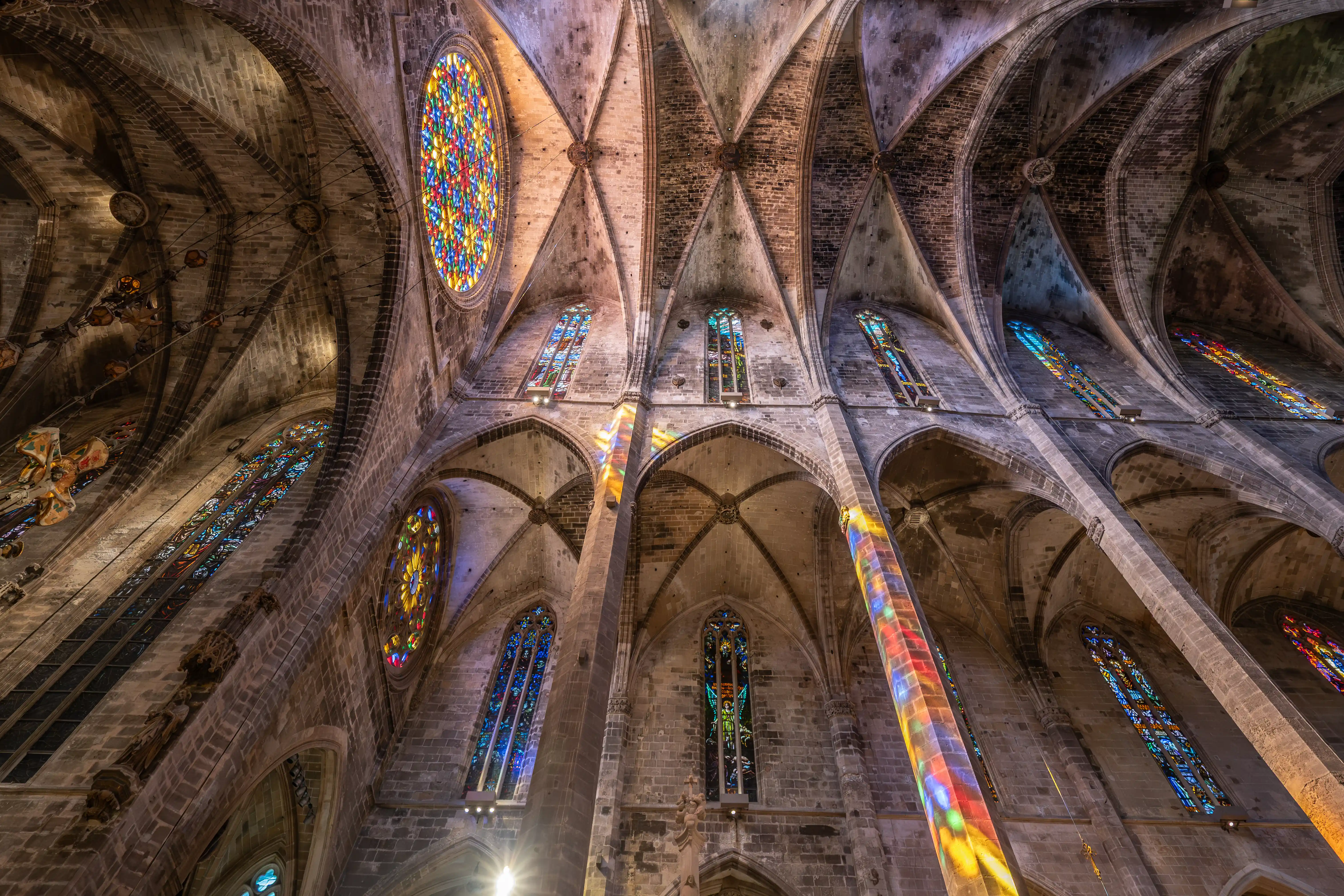 Interior of a Gothic cathedral with ornate vaulted ceilings and stone pillars, illuminated by colourful light from stained-glass windows.