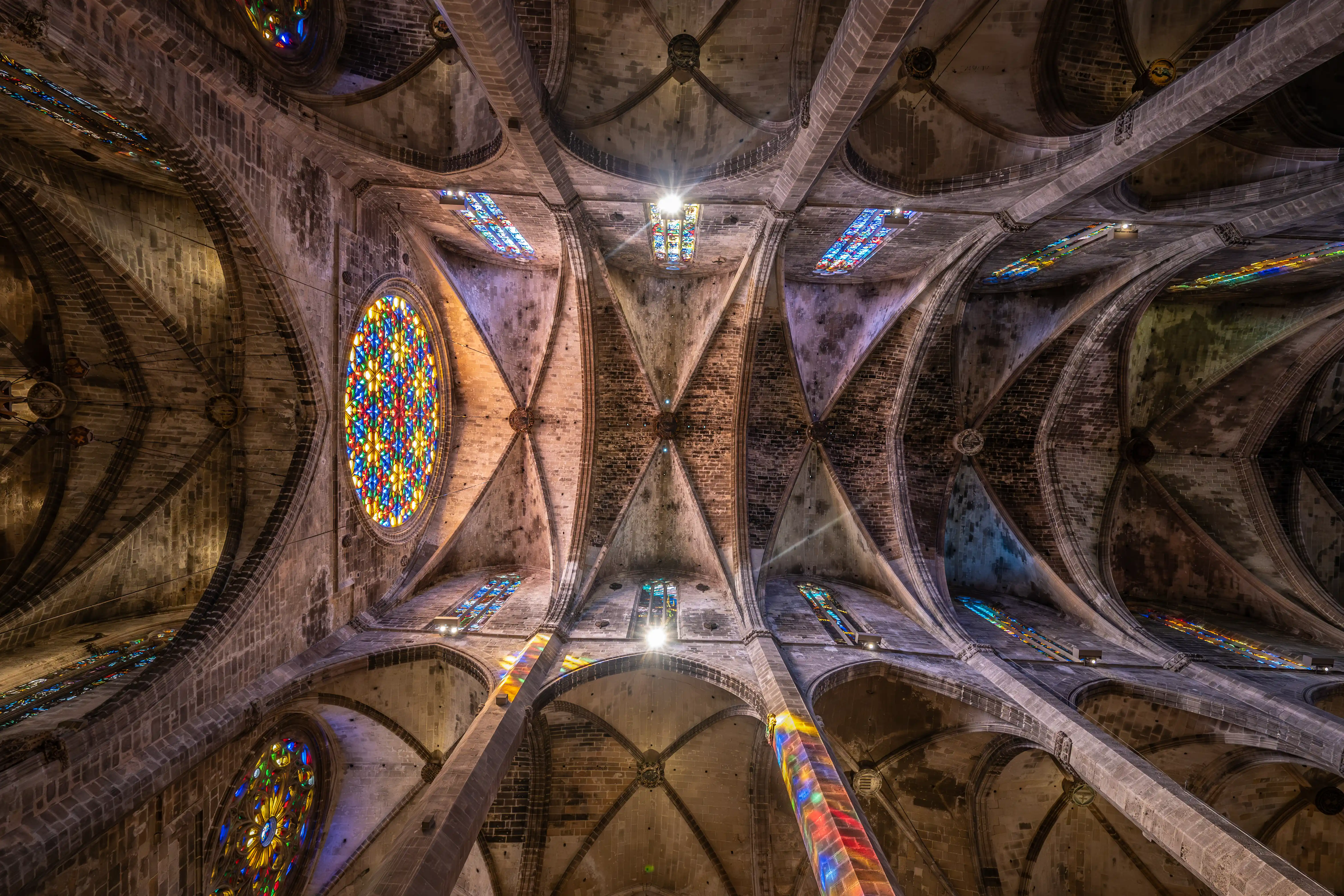  Wide-angle view looking upwards at the ornate vaulted ceiling of a Gothic cathedral, featuring colourful stained-glass windows and sunbeams illuminating the stone arches and brickwork.