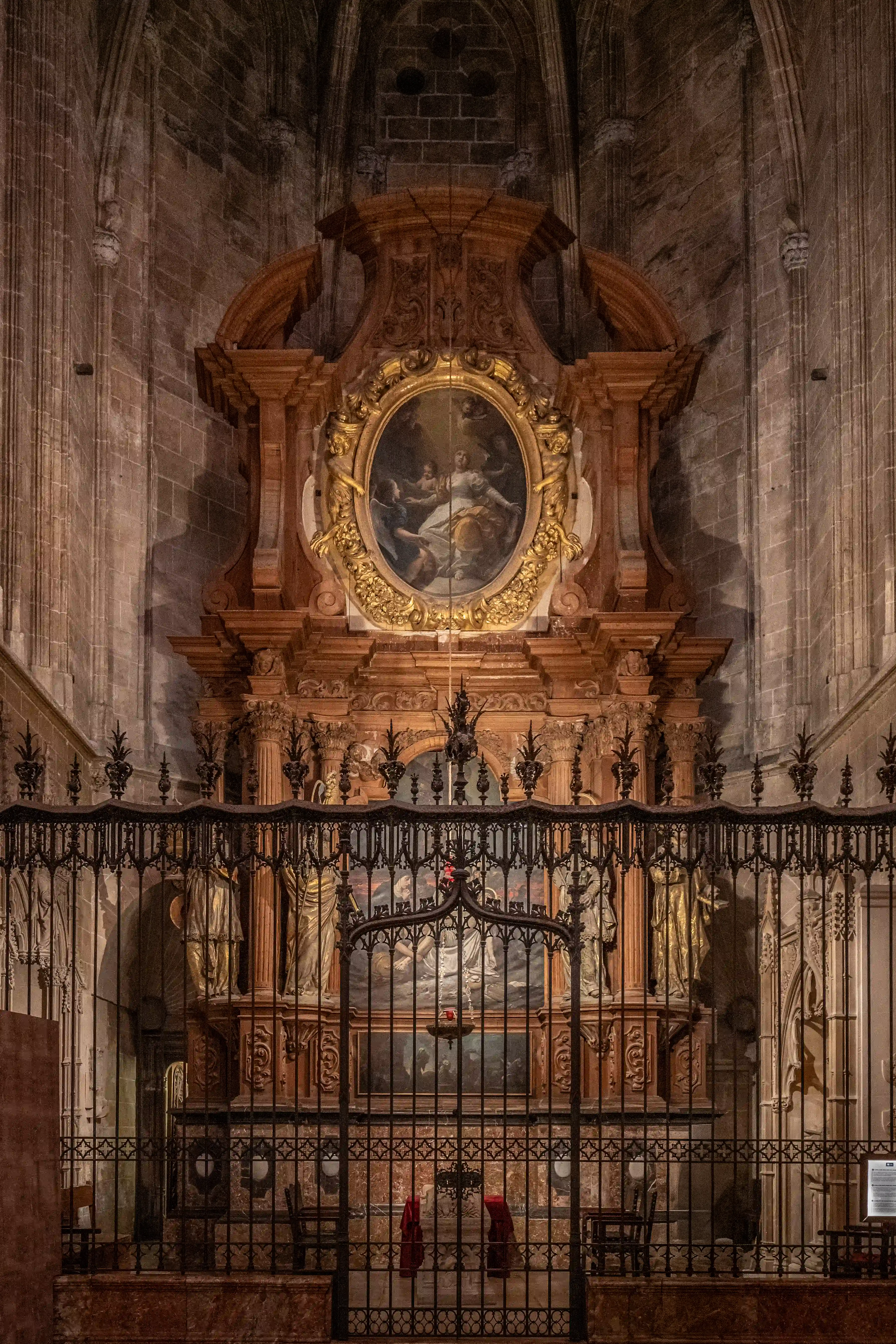 A richly decorated altar within a cathedral, featuring a central oval painting of religious figures and ornate wooden carvings, partially enclosed by a wrought iron screen that reveals further sculptures and paintings behind.