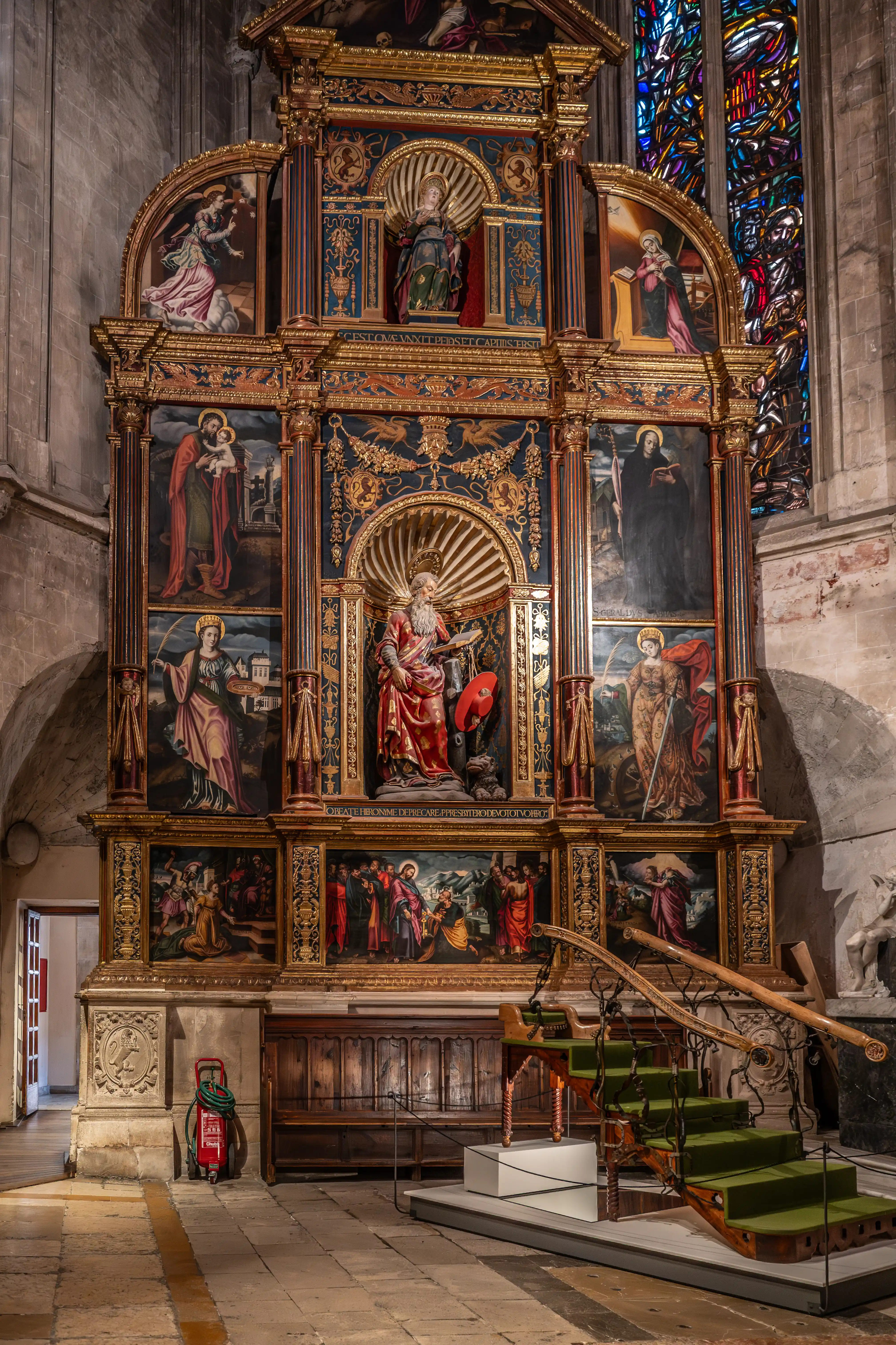 A large, ornate, gold and dark blue altarpiece in a church, displaying numerous religious paintings of saints and biblical scenes, a central sculpture of a bearded man, and intricate decorative details. Stained glass is visible to the right, and a modern green staircase is in the foreground.
