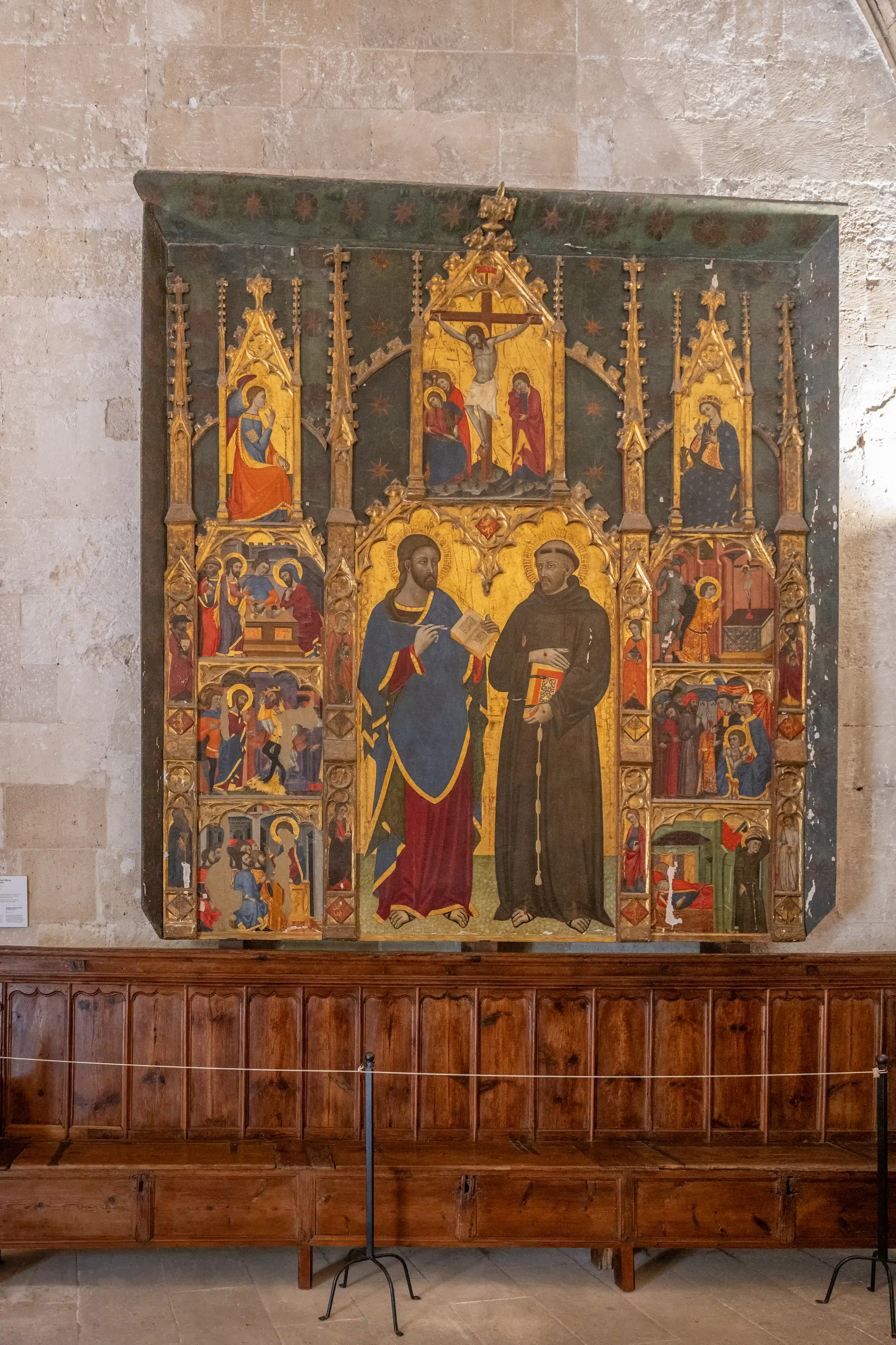 A large, gilded Gothic altarpiece featuring multiple painted panels of religious scenes, including a crucifixion and depictions of saints. In the centre, Christ is shown in conversation with Saint Francis. The altarpiece is mounted on a stone wall above antique wooden pews.