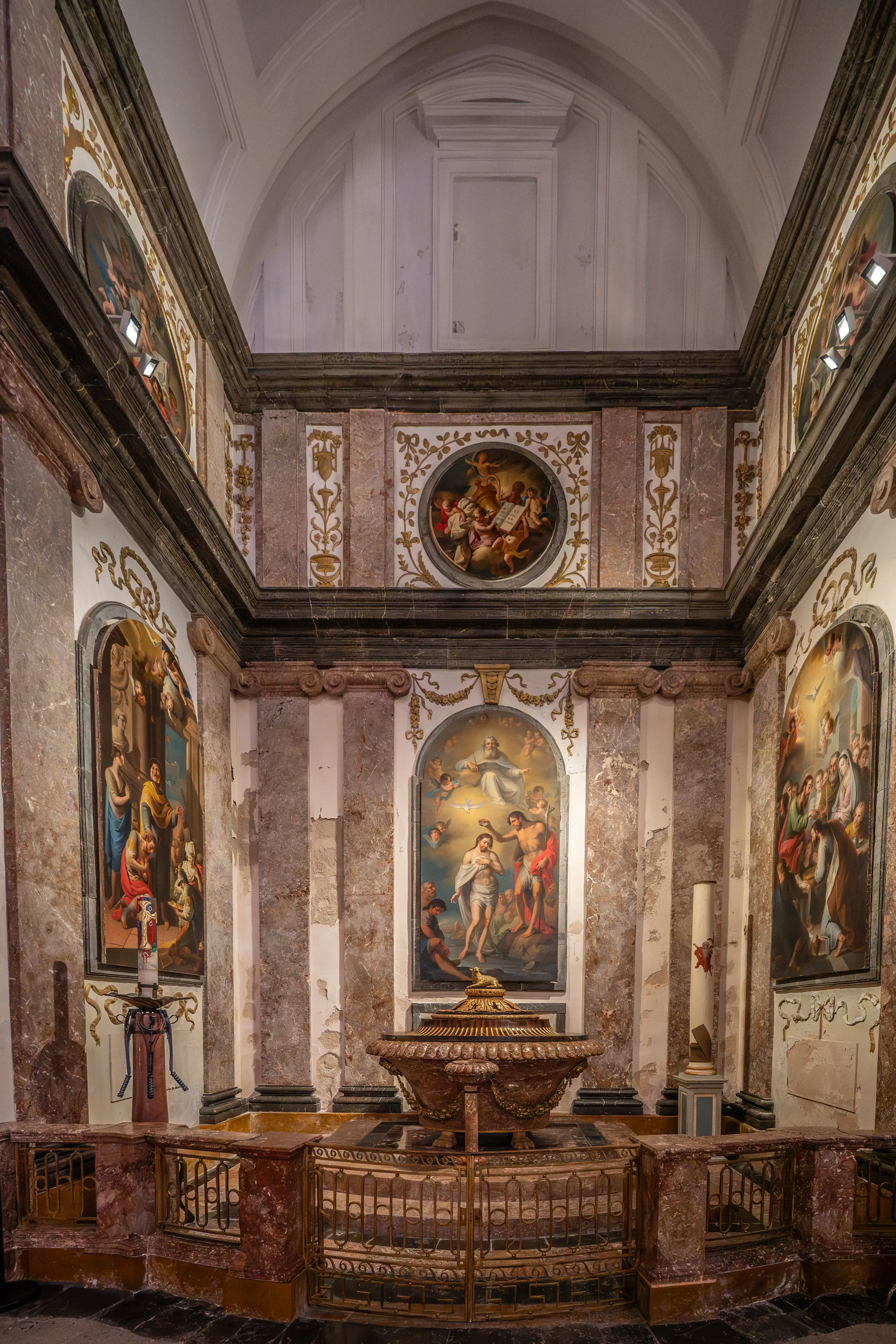 Interior view of a Baroque chapel with religious paintings, a central baptismal font, ornate metal railings, and marble-clad walls.