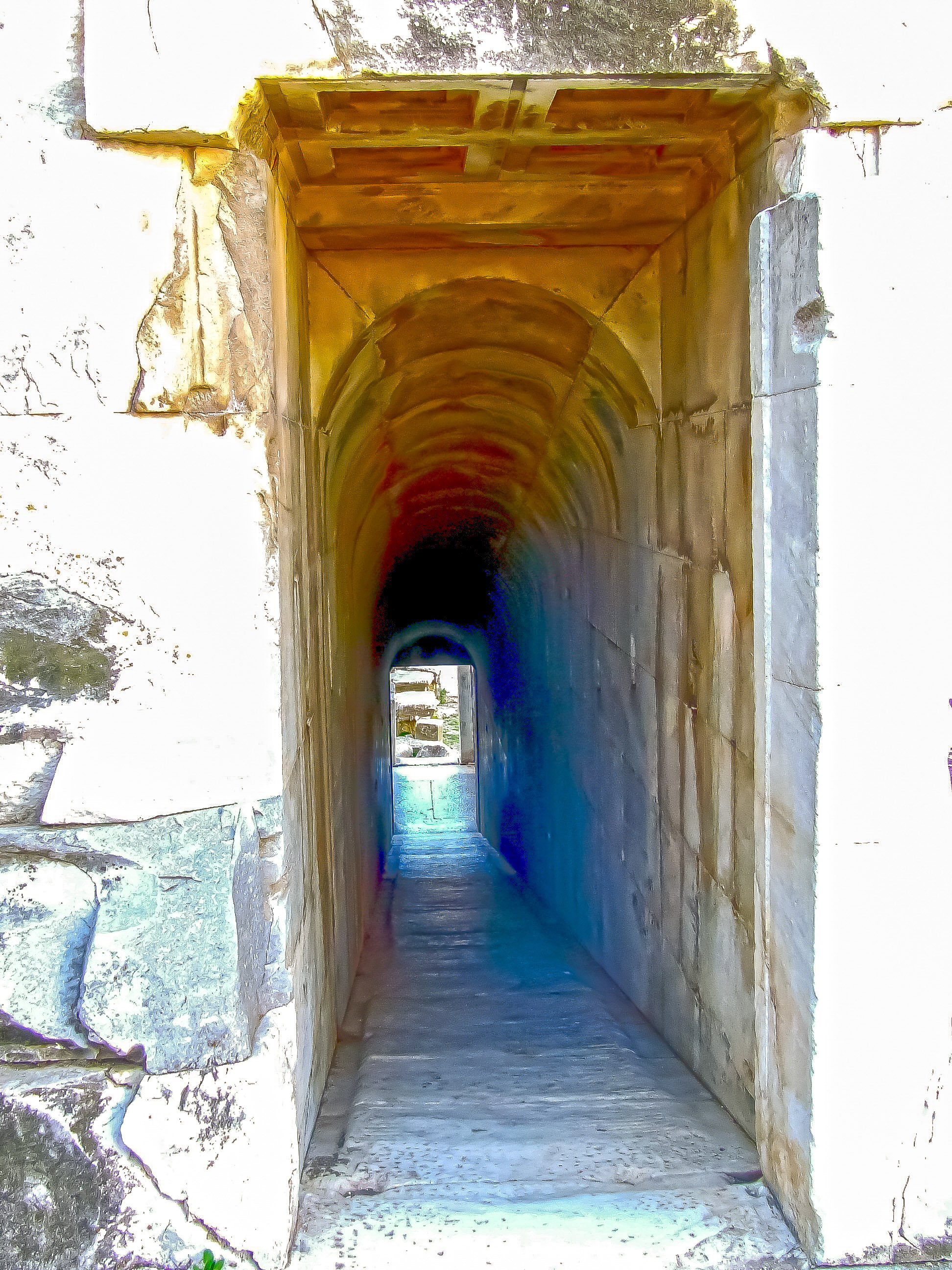 The image depicts a long, arched tunnel with a stone floor and walls. The tunnel appears to be well-lit, with sunlight creating a rainbow effect on the walls. The entrance to the tunnel is framed by a wooden structure, and the tunnel extends into the distance, leading to an open area at the end.