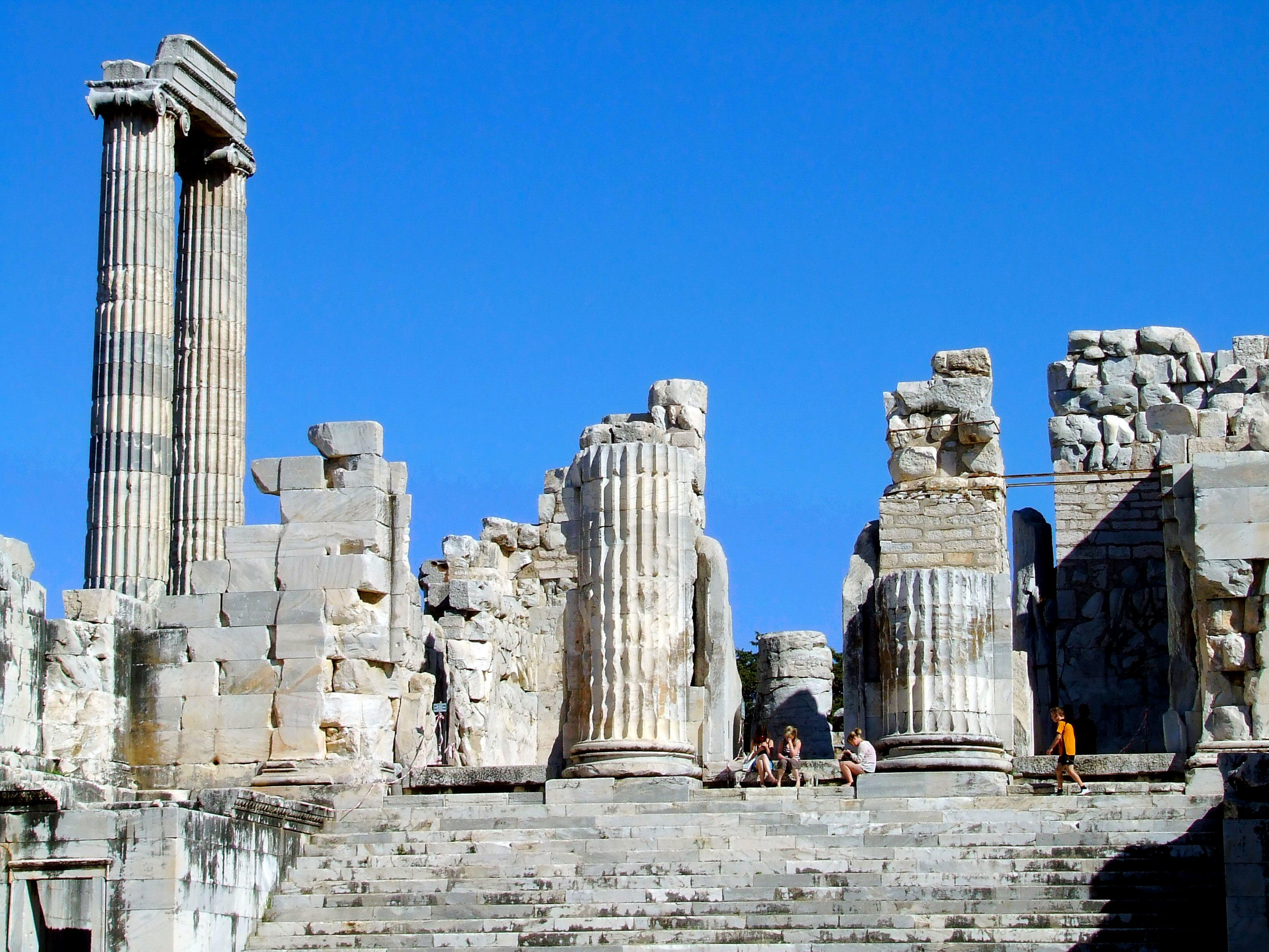 The image depicts the ancient ruins of a structure, likely a temple or significant building, characterized by tall, fluted columns and partially intact stone walls. The ruins are set against a clear blue sky, suggesting a sunny day. Several people are present, some sitting on the steps and others standing, indicating that the site is a tourist attraction. The overall scene conveys a sense of historical significance and architectural grandeur.