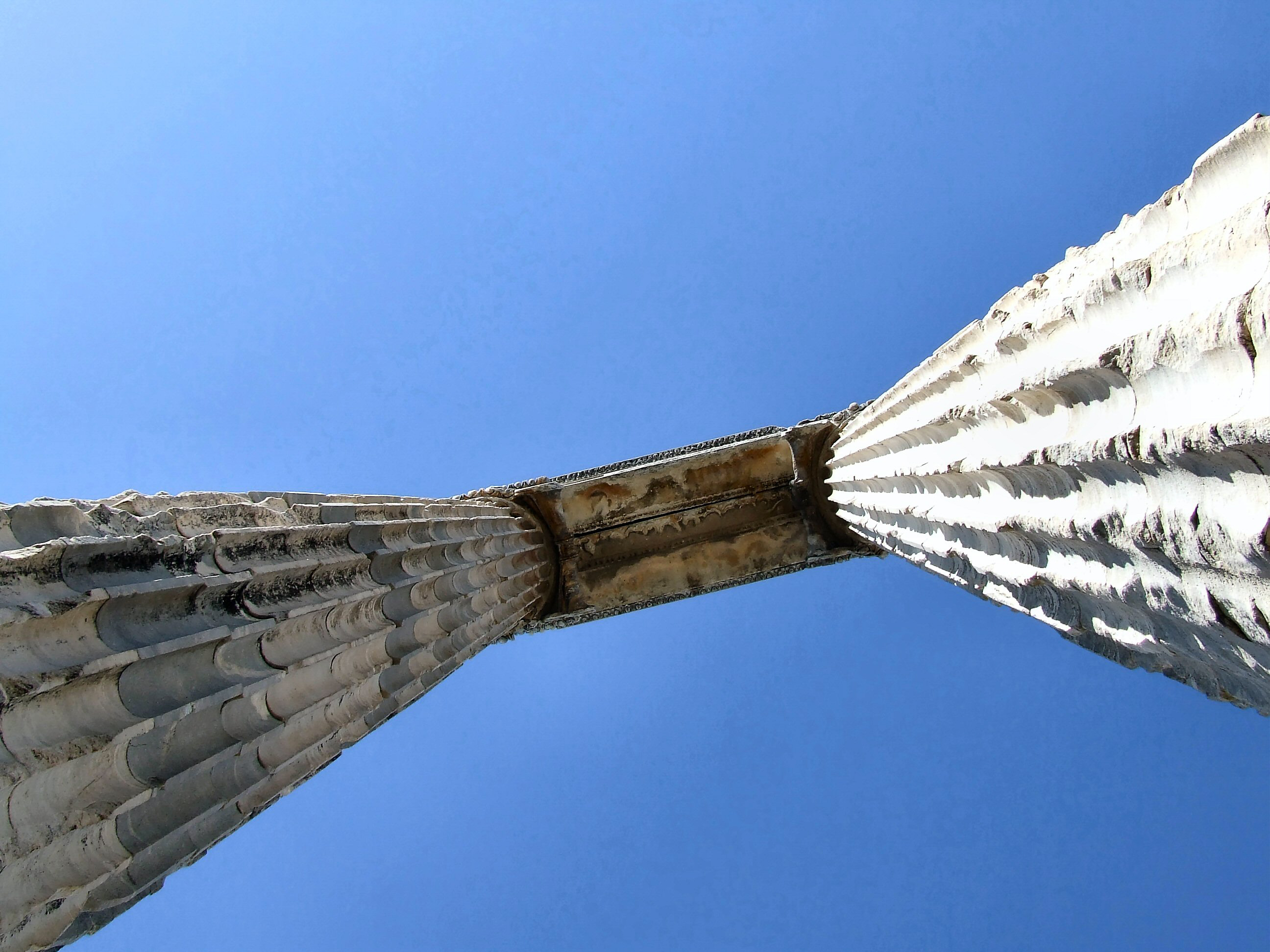 The image depicts a close-up view of a large, white, ribbed structure against a clear blue sky. The structure appears to be part of a large architectural or artistic installation, possibly a sculpture or a bridge. The ribs are arranged in a symmetrical pattern, converging towards a central point. The surface of the ribs shows signs of weathering and discoloration, indicating that the structure has been exposed to the elements for some time.