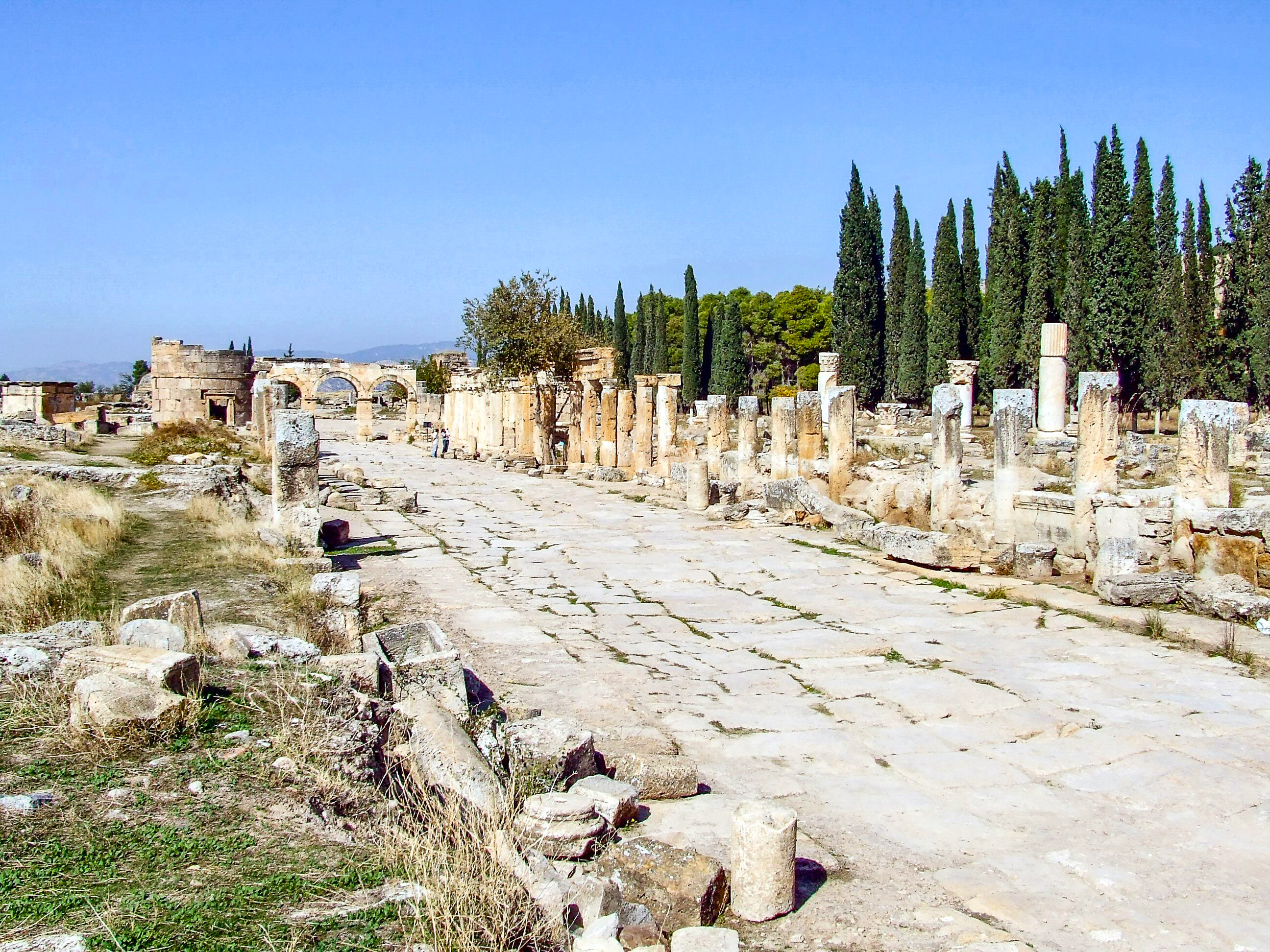 The image depicts an ancient archaeological site with a stone-paved pathway flanked by the remnants of stone columns and structures. The site is surrounded by tall cypress trees and other greenery, indicating a historical ruin possibly from a Roman or Greek era. The clear blue sky suggests a sunny day, and the overall scene conveys a sense of historical significance and tranquility.