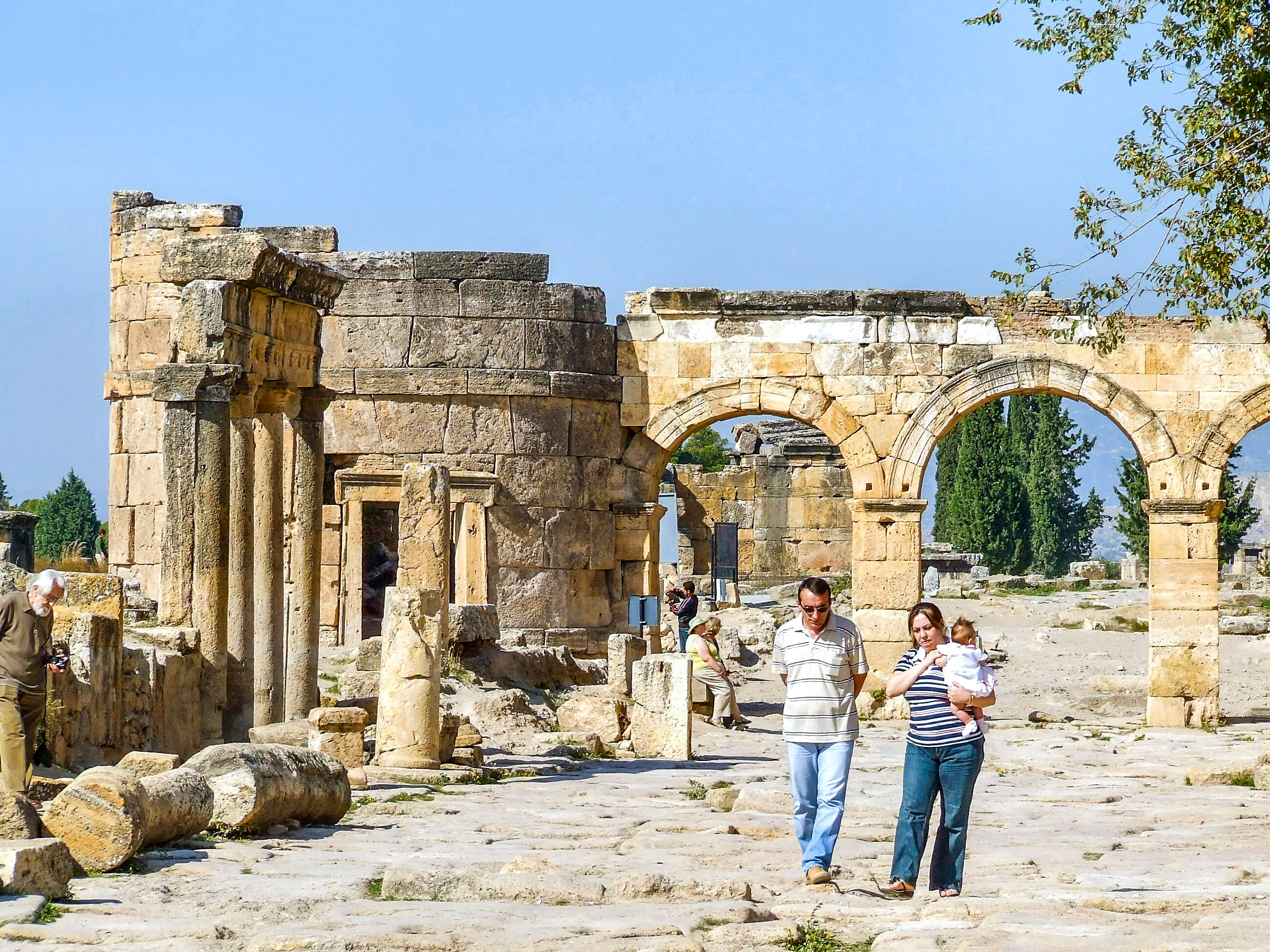 The image depicts an ancient archaeological site with stone ruins and columns. Several people, including a couple with a baby, are walking and exploring the site. The weather appears to be clear and sunny, and the surroundings include some greenery and trees.