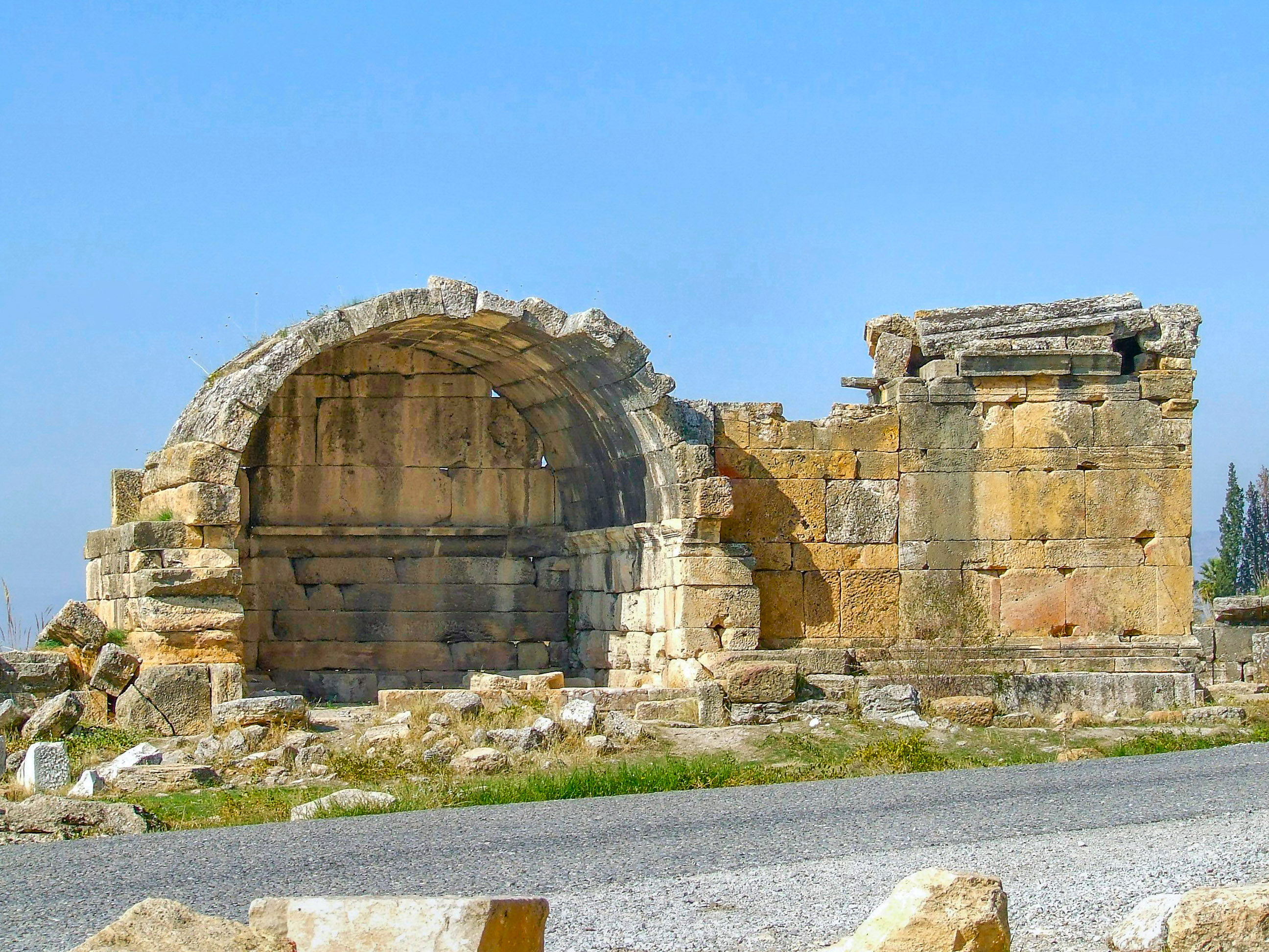 The image depicts an ancient stone structure with a prominent archway, likely part of a historical or archaeological site. The structure is made of large, weathered stones and appears to be in a state of ruin, with some parts of the walls and archway missing or crumbling. The ground around the structure is uneven and strewn with additional stones and debris, indicating it has not been maintained or restored recently. The sky is clear and blue, suggesting it is a sunny day. The overall scene conveys a sense of antiquity and historical significance.