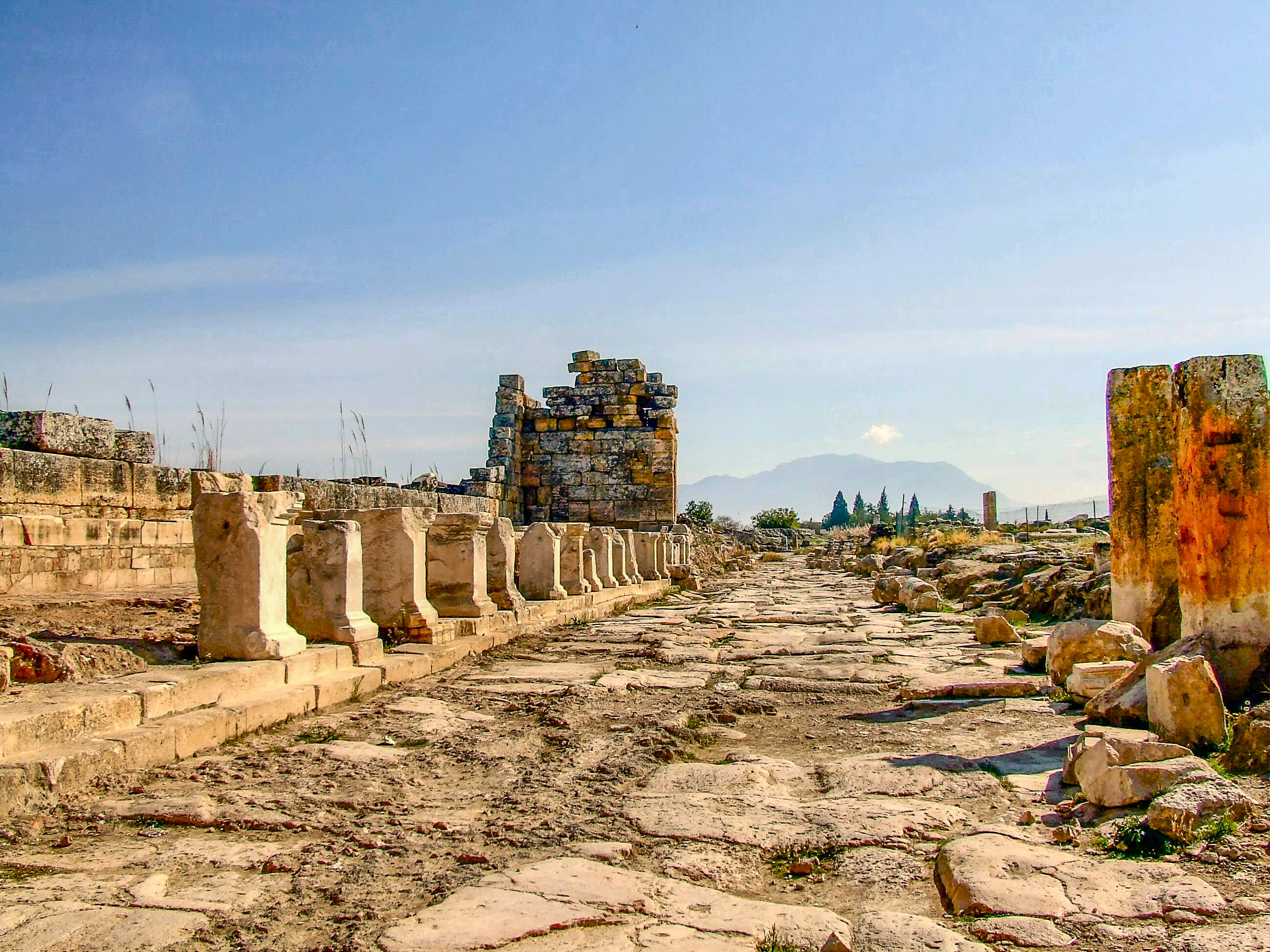 The image depicts the ruins of an ancient structure, likely a historical or archaeological site. The scene features old stone walls and columns, some of which are partially standing while others are in ruins. The ground is uneven with scattered stones and patches of dirt. In the background, there are distant mountains and a clear blue sky, suggesting a dry and possibly Mediterranean climate. The overall atmosphere is one of antiquity and historical significance.