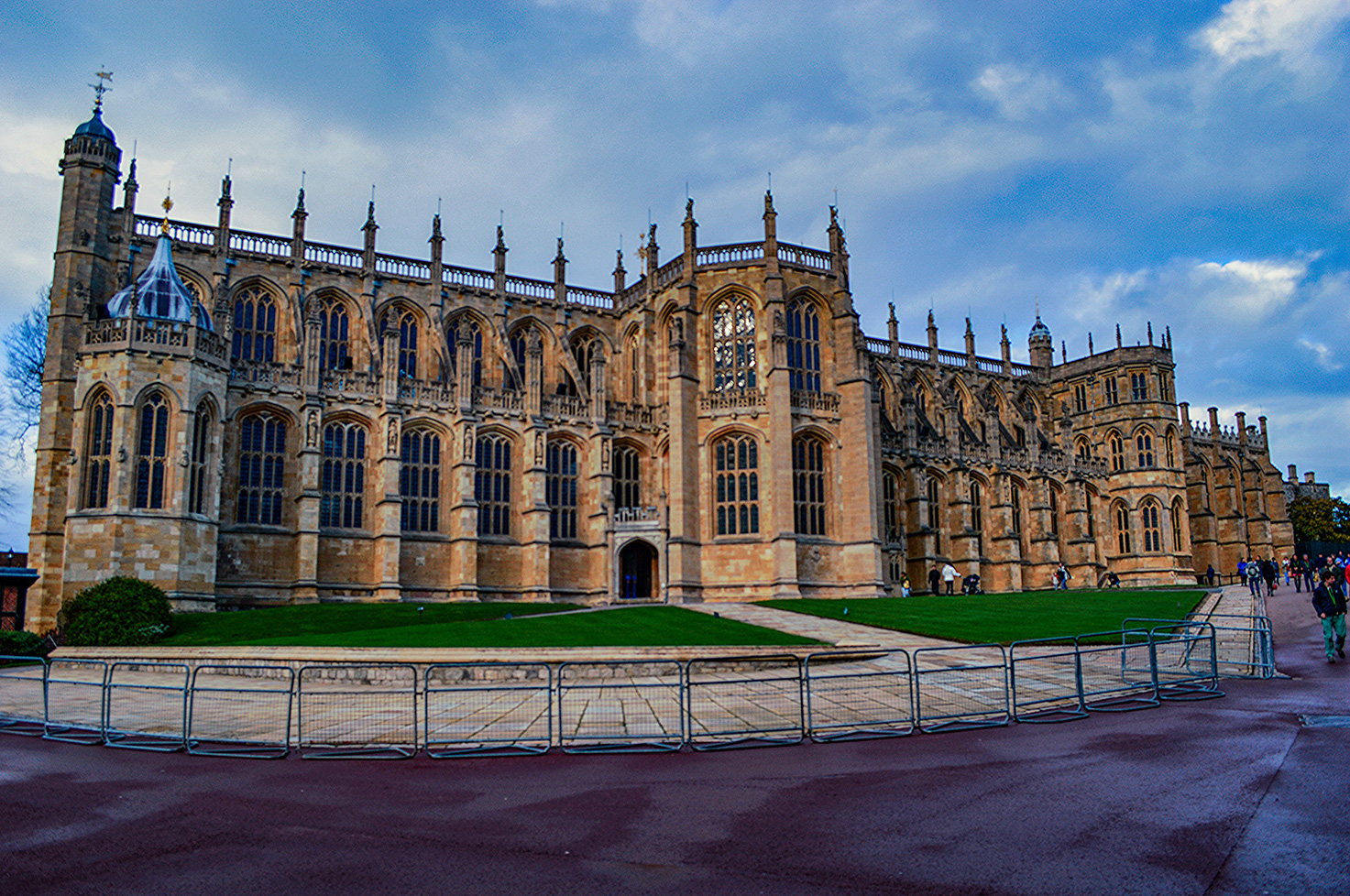 The image depicts a large, historic Gothic-style building with intricate architectural details, including pointed arches, ribbed vaults, and flying buttresses. The structure features numerous tall, narrow windows with elaborate tracery and large stained glass windows. The building is constructed from stone and has multiple towers and spires, adding to its grandeur. In front of the building, there is a well-maintained lawn and a pathway with metal barriers, suggesting it is a significant and possibly protected site. The sky above is partly cloudy, and there are a few people visible near the building, indicating it might be a tourist attraction or a place of historical importance.
