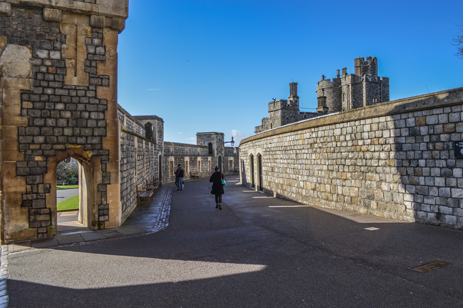 The image depicts a stone walkway within a castle or fortress. The walkway is lined with high stone walls and arches, leading towards a larger stone structure in the background. The sky is clear and blue, and there are a few people walking along the path. The architecture suggests a historical or medieval setting.