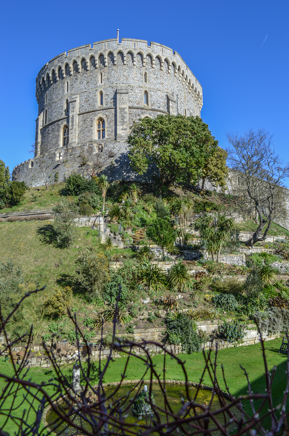The image depicts a historic, round stone tower situated on a hill. The tower is surrounded by lush greenery, including various trees and plants. There is a well-maintained garden with a small pond in the foreground. The sky is clear and blue, suggesting a bright, sunny day. The overall scene conveys a sense of tranquility and historical significance.