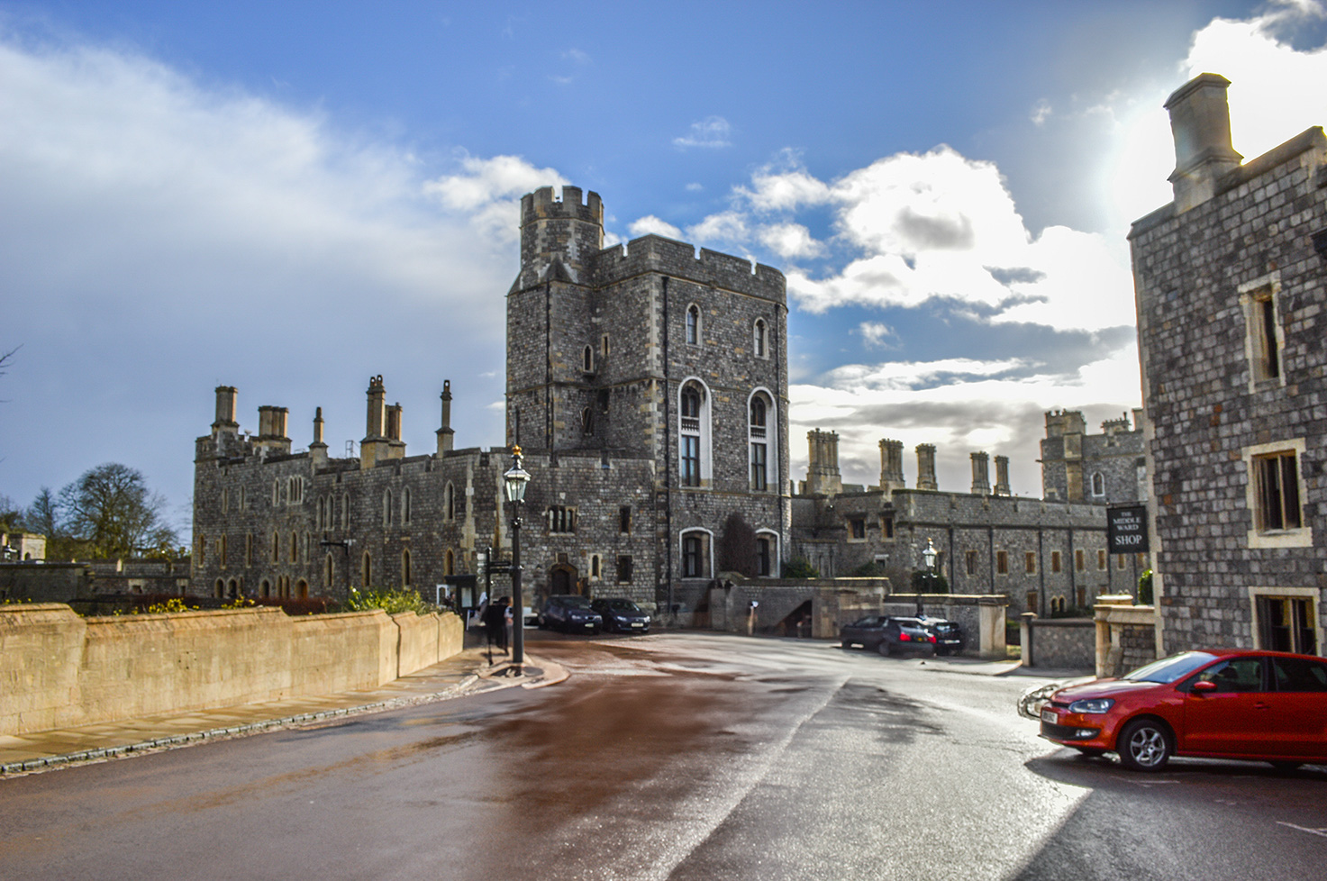 The image depicts a historic castle-like building with stone walls and multiple towers. The structure has a medieval architectural style, featuring arched windows and chimneys. The scene includes a street in front of the building with parked cars and a streetlamp. The sky is partly cloudy, and the overall atmosphere suggests a blend of historical and modern elements.