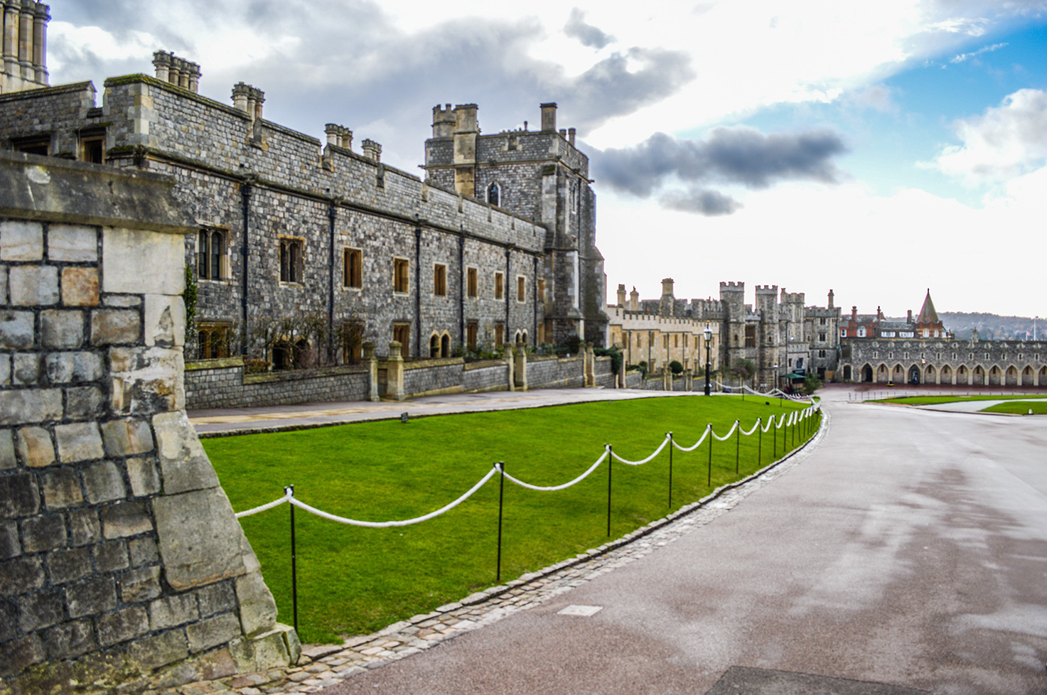 The image depicts a large, historic castle with stone walls and multiple towers. The castle is surrounded by a well-maintained green lawn and a paved pathway. The sky above is partly cloudy, and the overall scene suggests a serene and majestic atmosphere.