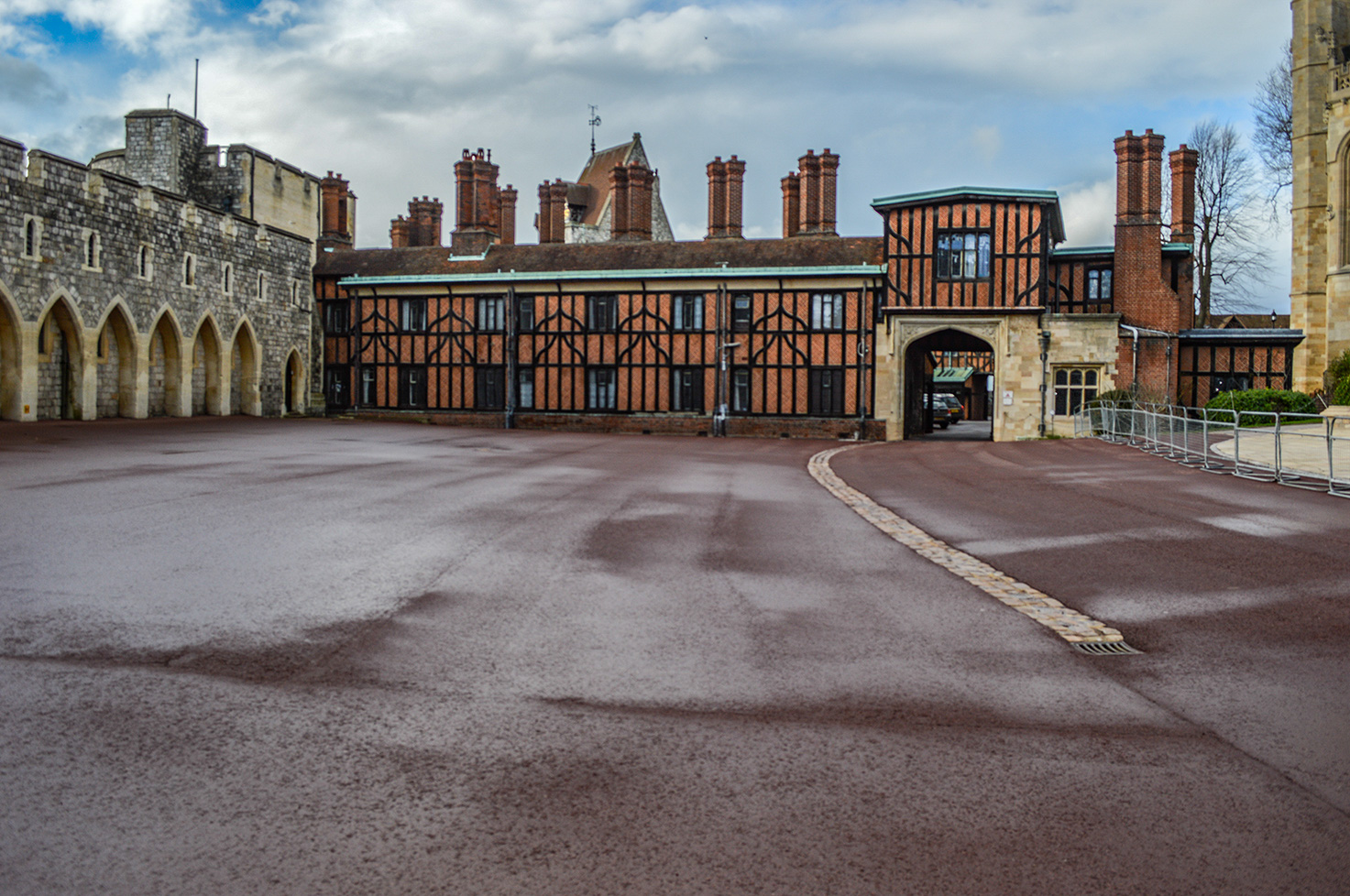 The image depicts a historic building with a large courtyard. The structure features a combination of stone and timber-framed architecture, with multiple chimneys and a distinctive arched entrance. The courtyard is paved and appears to be part of a larger complex, possibly a castle or a manor.