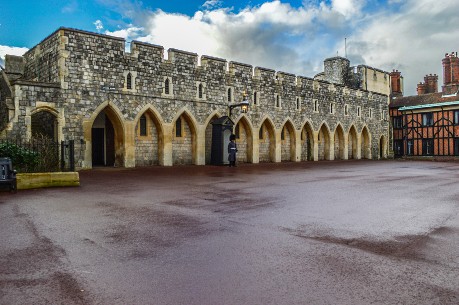The image depicts a historic stone building with a series of pointed arches and a guard standing at attention in front of it. The structure appears to be part of a castle or fortress, characterized by its robust stone walls and battlements. The sky is partly cloudy, and the ground is wet, suggesting recent rain. The scene conveys a sense of history and formality.