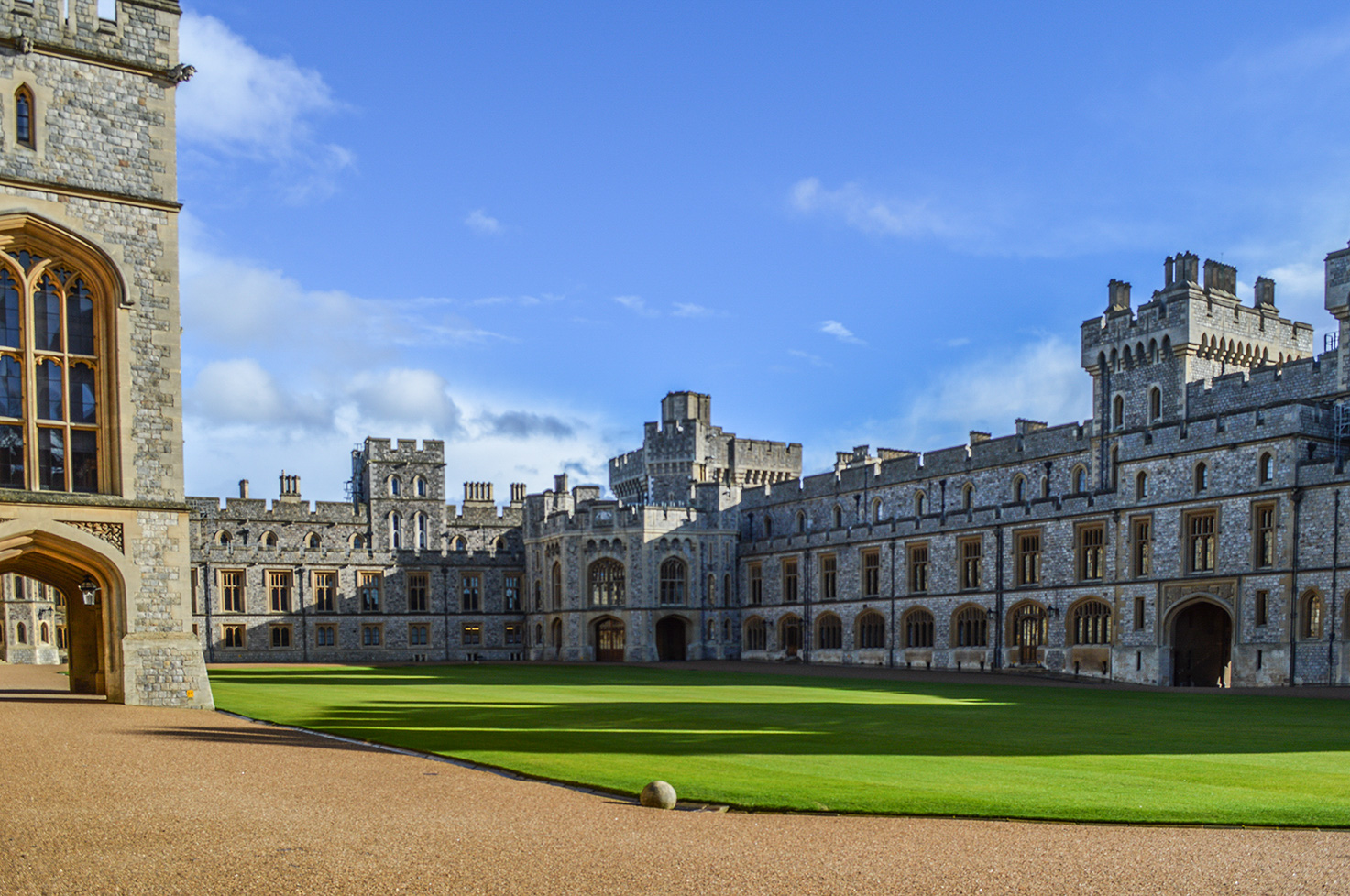 The image depicts a grand, historic castle with a large courtyard. The castle features tall, stone towers and walls with numerous windows and arched doorways. The architecture is Gothic in style, characterized by pointed arches and detailed stonework. The courtyard is paved with a mix of grass and gravel, and the sky above is clear with a few scattered clouds.
