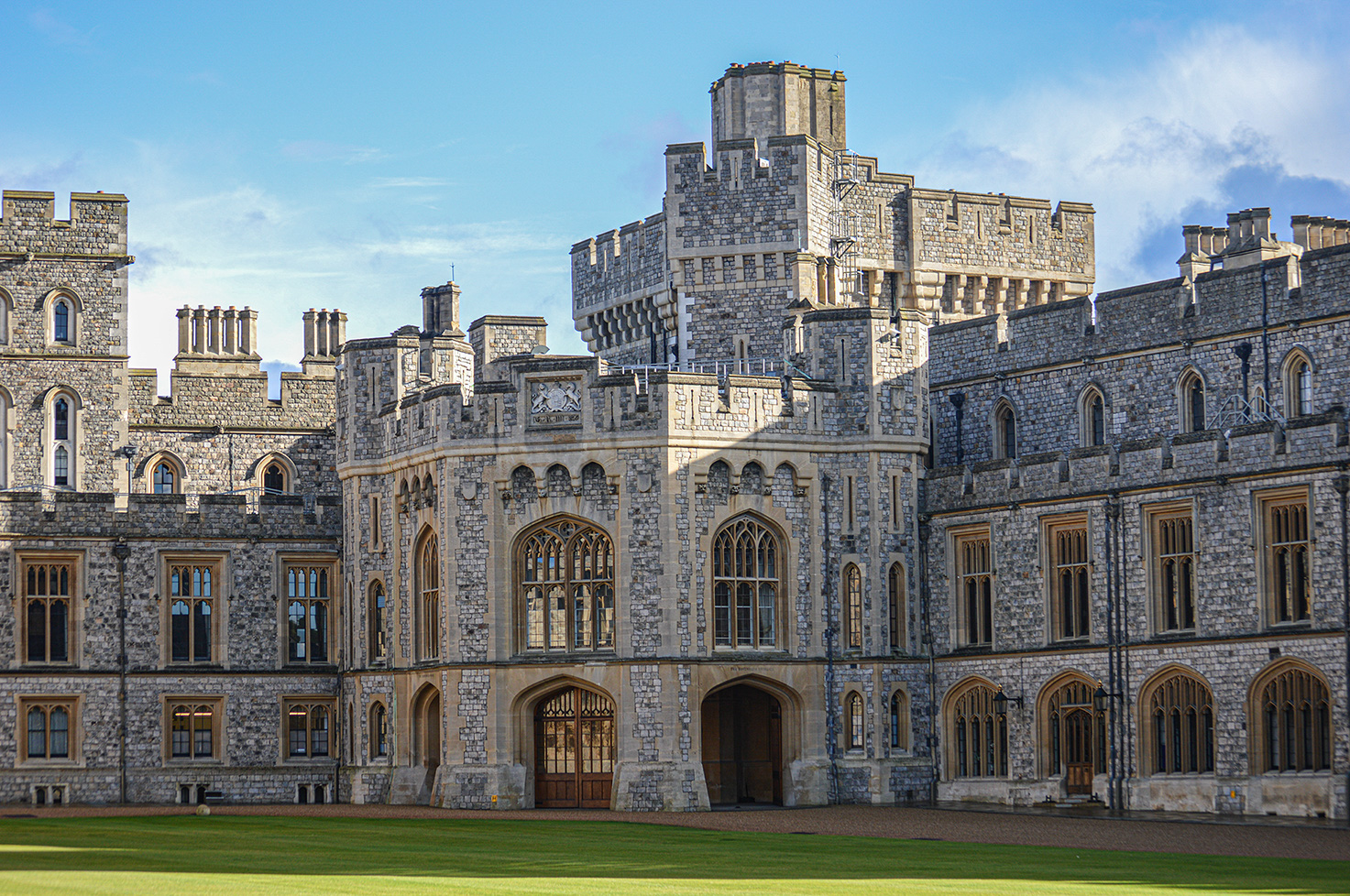 The image depicts Windsor Castle, a historic royal residence located in Windsor, England. The castle, constructed primarily of stone, features a mix of Gothic and Romanesque architectural styles. It has multiple towers, turrets, and large windows with intricate designs. The foreground shows a well-maintained lawn leading up to the castle's entrance. The sky above is clear with a few scattered clouds, adding to the picturesque scene.