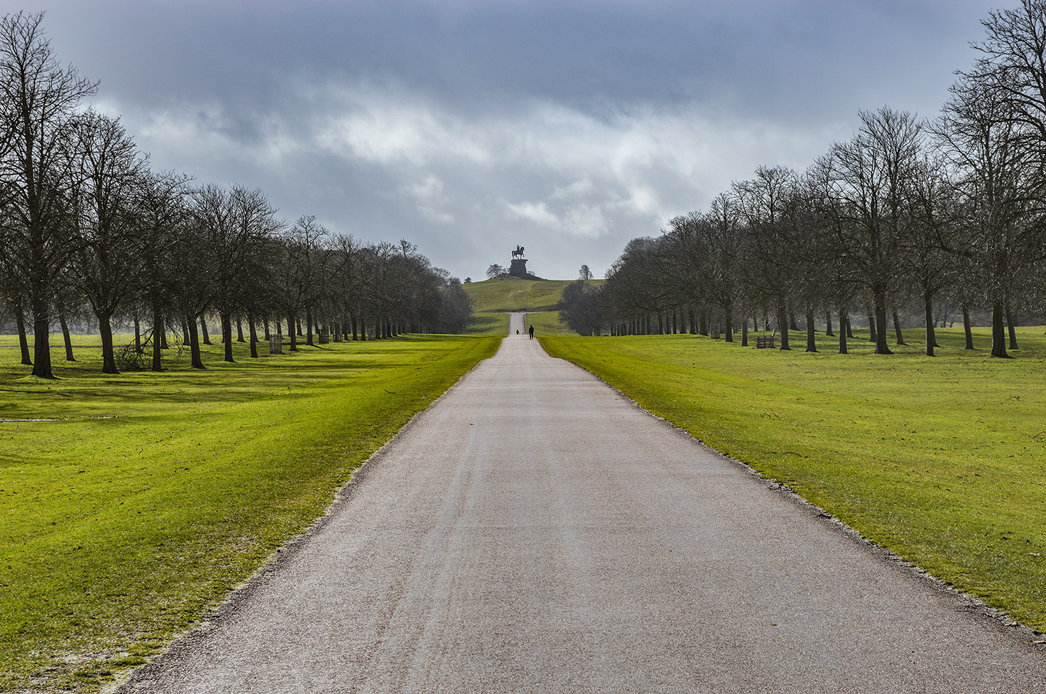 The image depicts a long, straight path leading to a hill topped with a statue. The path is flanked by rows of trees on both sides, creating a symmetrical and picturesque scene. The sky is overcast, adding a dramatic effect to the landscape. The path appears to be well-maintained, and there is a person walking towards the statue in the distance.