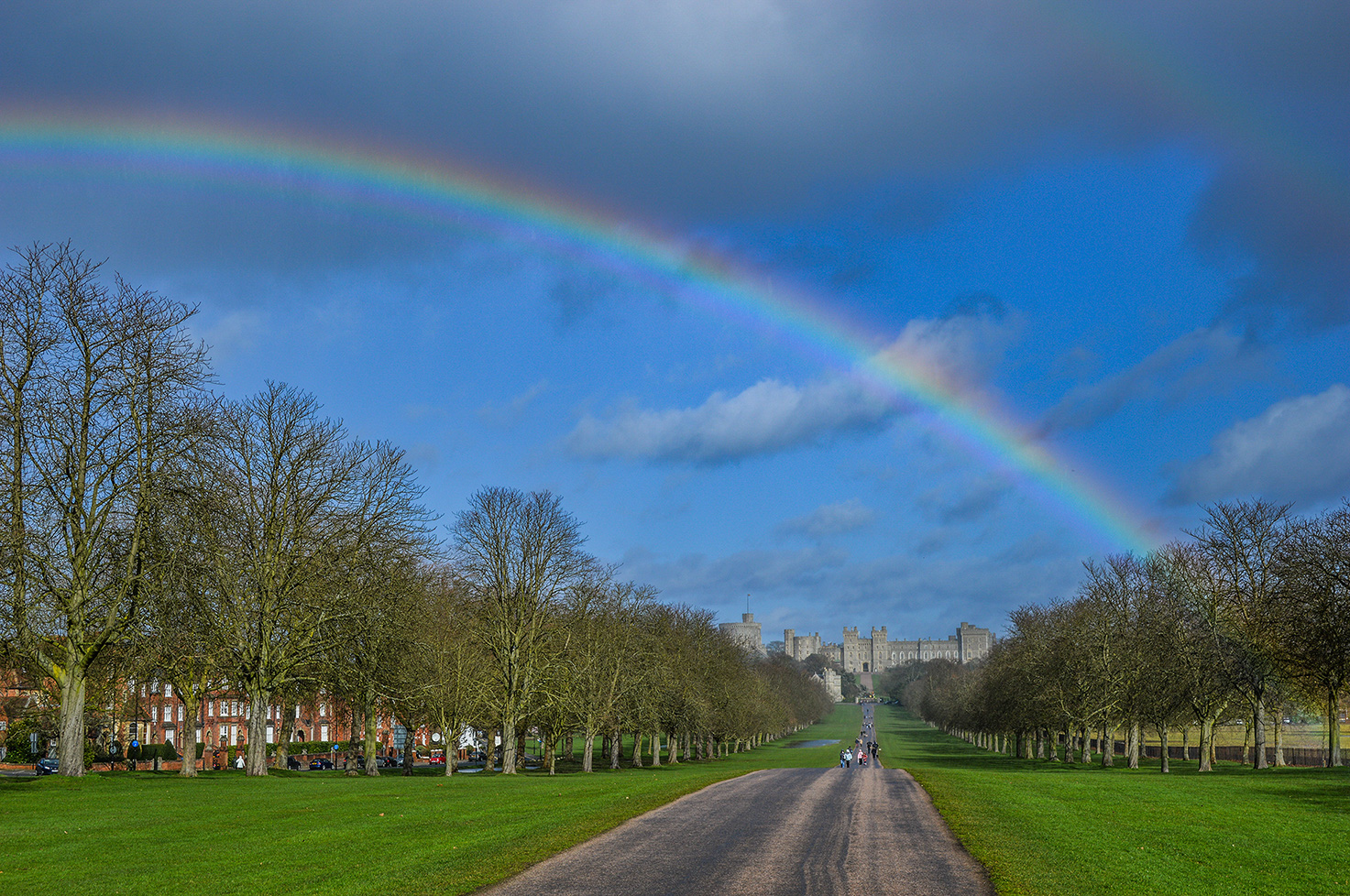 The image depicts a scenic view of Windsor Great Park with a prominent double rainbow arching across the sky. The park features a long, straight path flanked by leafless trees, leading towards Windsor Castle in the distance. The sky is partly cloudy with a mix of blue and grey tones. On the left side of the path, there are residential buildings, and the right side is bordered by expansive green lawns. A few people can be seen walking or biking along the path, enjoying the picturesque landscape.