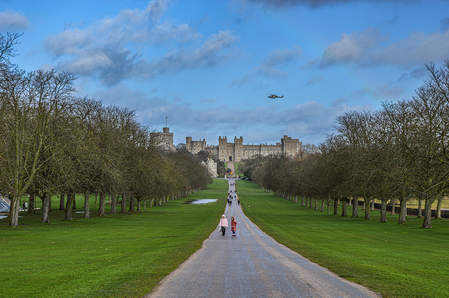 The image depicts a scenic view of Windsor Castle, a historic royal residence in England. The path leading to the castle is lined with trees and people walking towards the castle. The sky is partly cloudy, and a helicopter is seen flying above the castle.