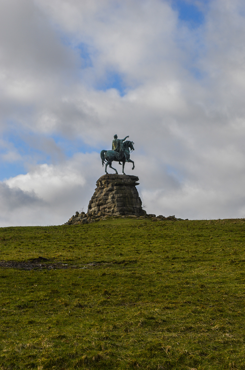 The image shows a statue of a man on horseback positioned on a large stone pedestal situated on a grassy hill under a partly cloudy sky.