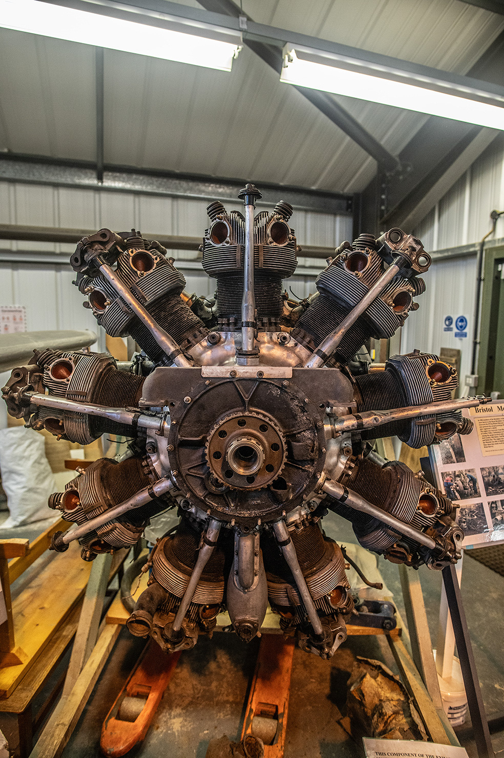 The image shows a radial engine, likely from an aircraft, displayed in a museum setting. The engine has multiple cylinders arranged radially around a central crankcase, with visible exhaust pipes and other components. The setting appears to be an indoor exhibit with informational placards nearby.