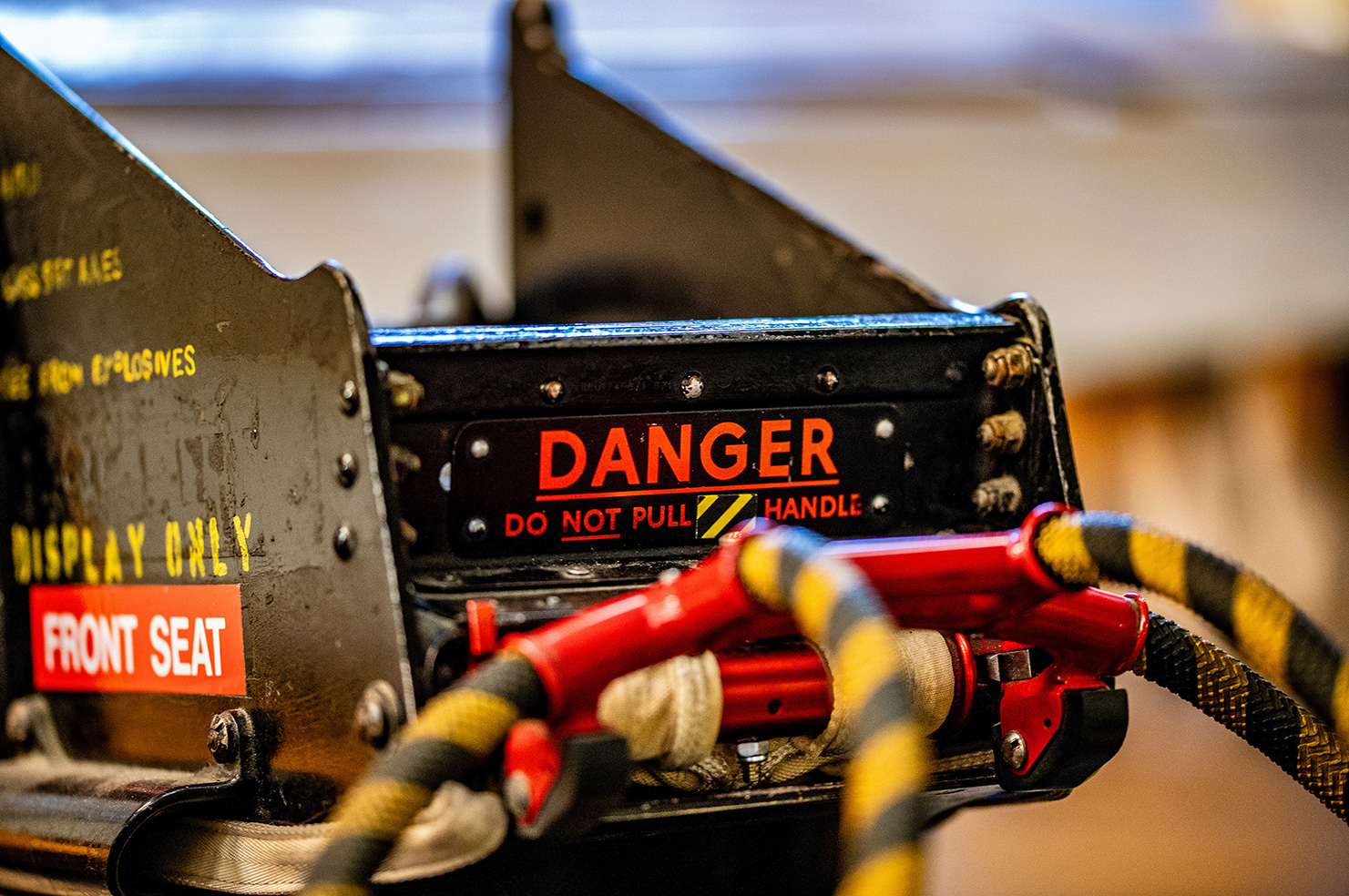The image shows a close-up of a mechanical device, likely part of an aircraft, with several warning labels. The most prominent label reads 'DANGER DO NOT PULL HANDLE,' indicating a critical safety instruction. Other labels include 'DISPLAY ONLY' and 'FRONT SEAT,' suggesting that this component is not to be operated and is meant for display purposes only. The device appears to be secured with straps and has a red handle, emphasizing the importance of not tampering with it.