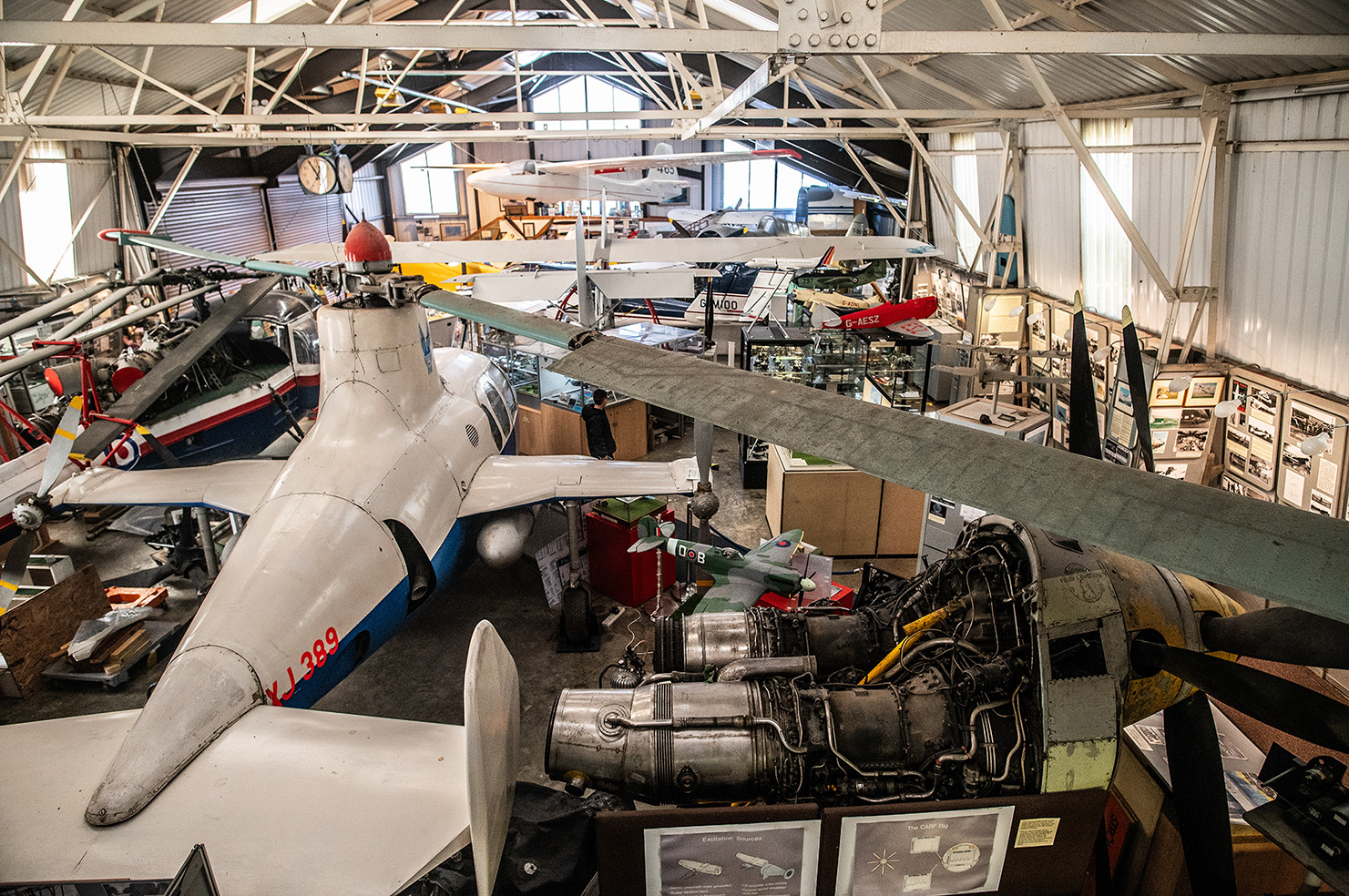 The image depicts the interior of an aviation museum, showcasing a variety of vintage aircraft and aircraft engines. The museum is housed in a large, well-lit hangar with a high ceiling. Several airplanes are on display, including a prominently featured aircraft in the foreground with a visible engine and propeller. The museum appears to have detailed informational displays about the exhibits, and there are various other aviation artifacts and memorabilia scattered throughout the space.
