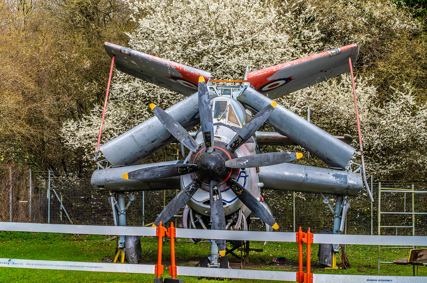 The image depicts an aircraft mounted on a stand for display. The aircraft is a vintage military plane, likely a fighter from the World War II era, as indicated by its design and markings. It is suspended in the air with its wings spread out, showcasing its propeller and cockpit. The plane is situated outdoors, surrounded by a fence and greenery, suggesting it is part of an outdoor museum or exhibition.