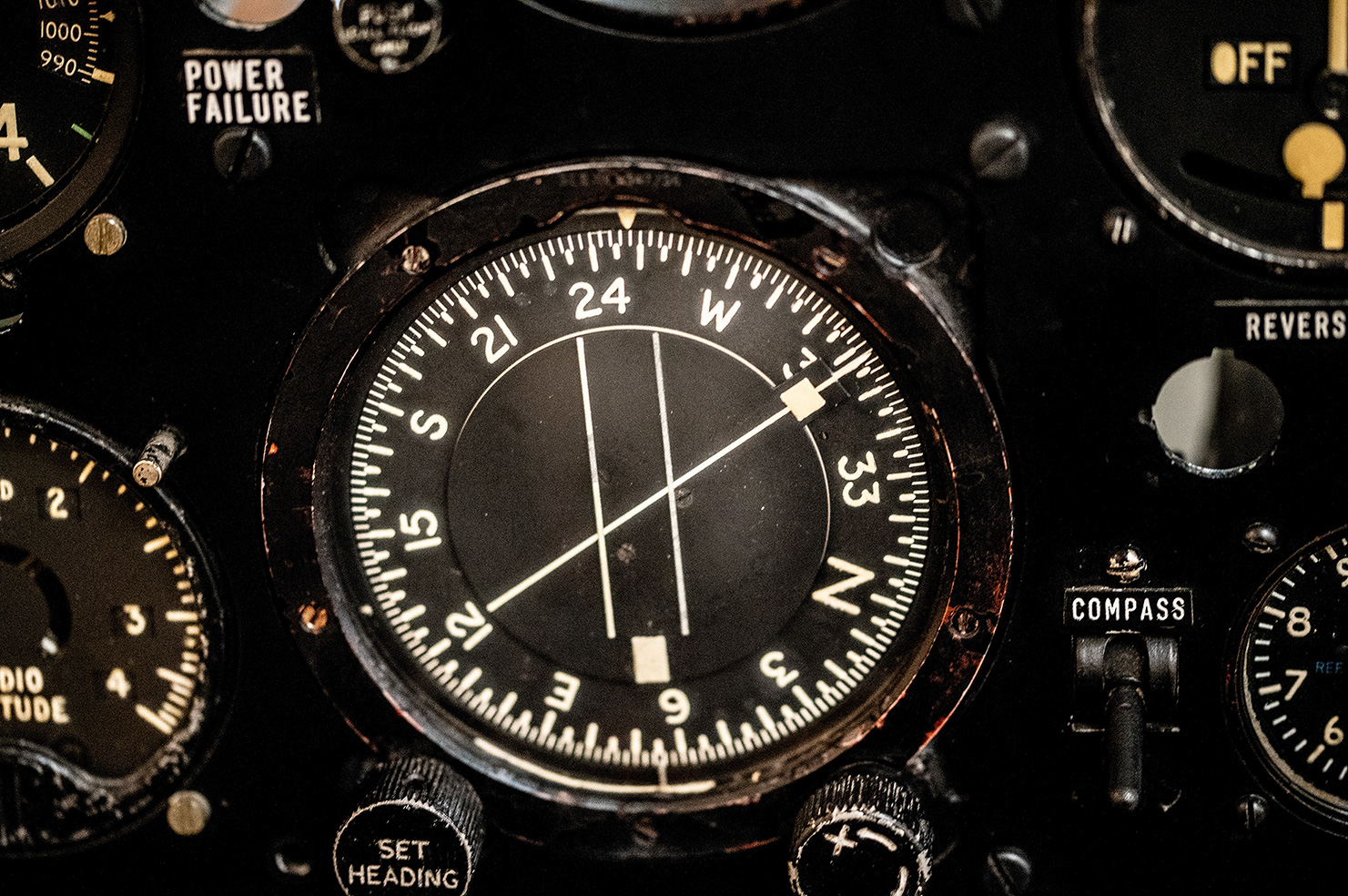 The image shows a close-up of an aircraft's compass and various other instruments on the control panel. The compass is prominently displayed in the center, with markings for cardinal directions (N, E, S, W) and degrees. Surrounding the compass are other instruments and labels such as 'POWER FAILURE,' 'OFF,' 'REVERS,' 'RADIO TUDE,' and 'SET HEADING.' The instruments appear to be part of an older aircraft's control panel, indicating navigation and communication tools used by the pilot.