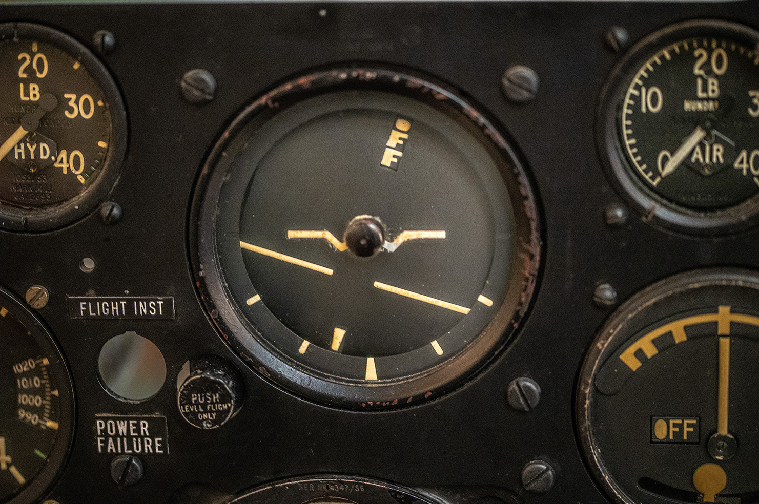 The image shows a close-up of an aircraft's instrument panel featuring several gauges and dials. The central gauge indicates the aircraft's attitude with a small airplane symbol and horizontal lines. Surrounding gauges measure hydraulic pressure, air pressure, and other critical flight parameters. Labels such as 'FLIGHT INST,' 'POWER FAILURE,' and 'PUSH TO TALK' are visible, indicating various functions and warnings. The overall appearance suggests a vintage or older aircraft cockpit design.