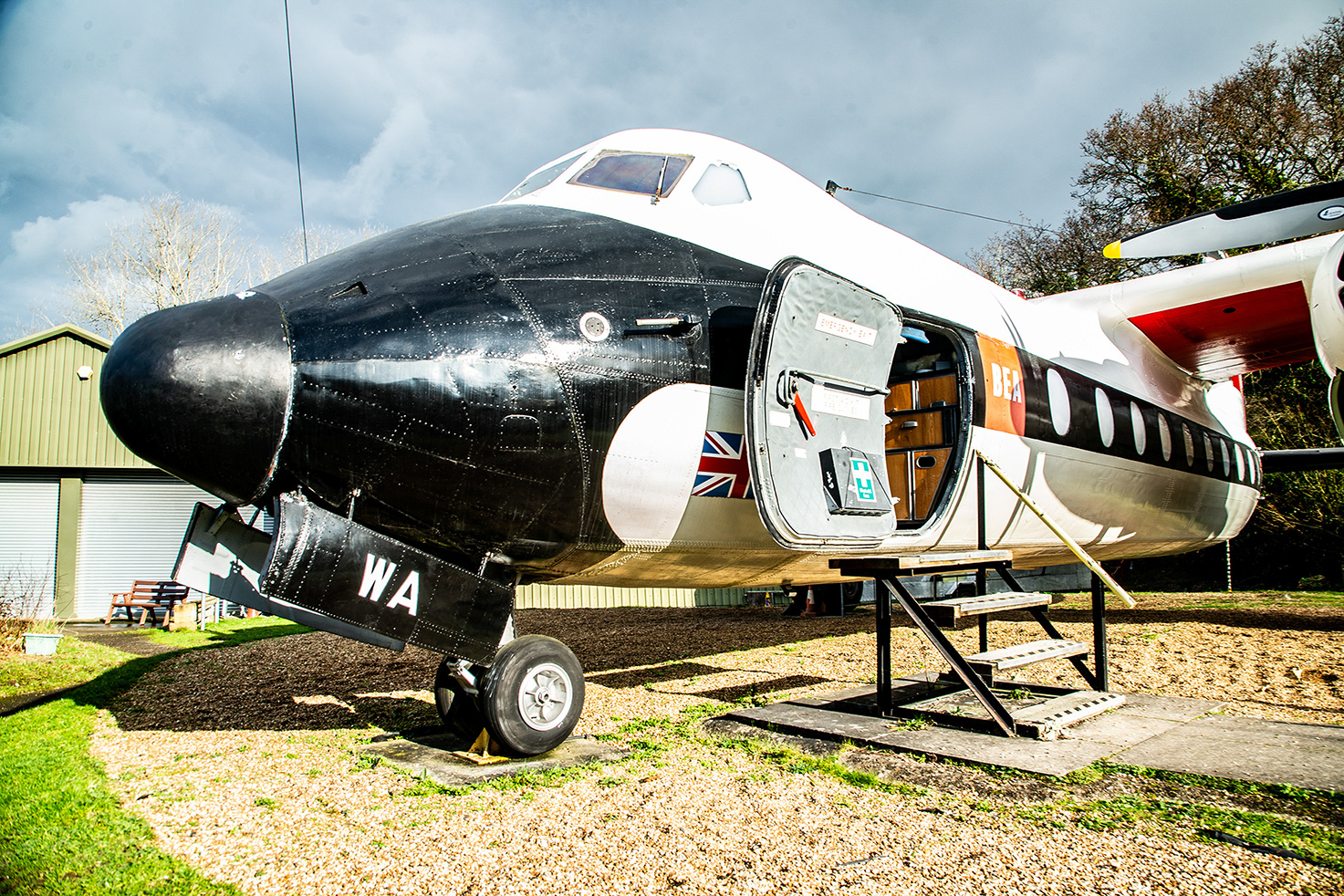 he image depicts a vintage aircraft with its door open, revealing a glimpse of the interior. The plane is parked on a grassy area with a small set of stairs leading up to the entrance. The aircraft has a distinctive black and white color scheme with some red and yellow accents. The tail of the plane has the letters 'WA' painted on it. The setting appears to be an outdoor display, possibly at a museum or an airfield.