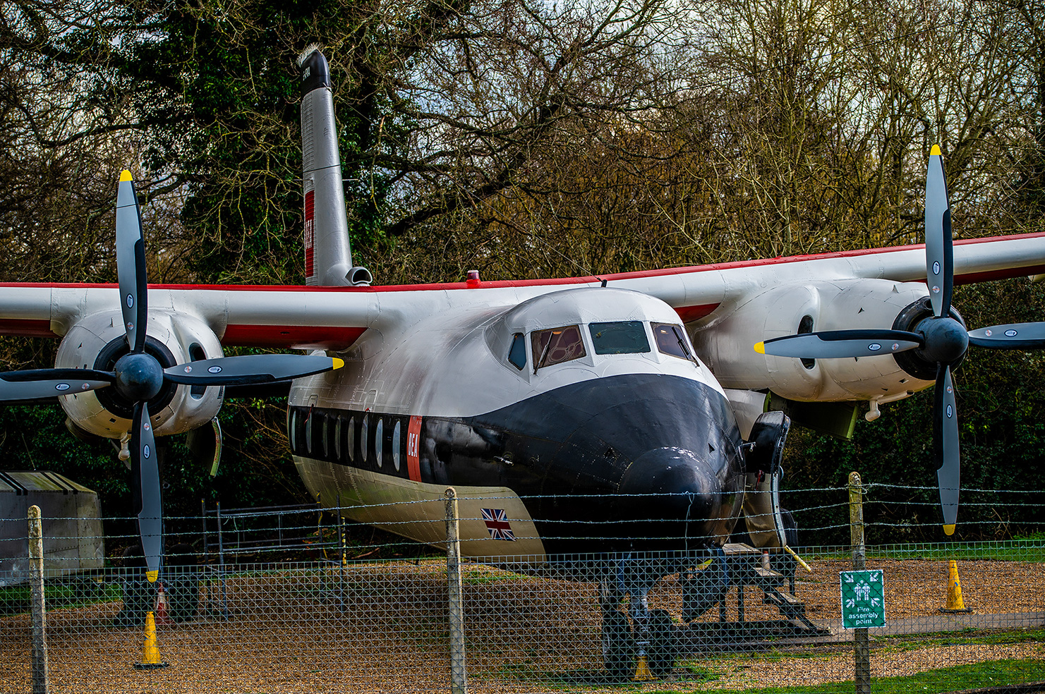 The image shows a large, vintage aircraft with four propellers, each with four blades. The aircraft is painted in a camouflage pattern of black, green, and light brown. It has a distinctive red and white stripe running along its side. The plane is parked on a grassy area enclosed by a chain-link fence. The background features trees with bare branches, suggesting it might be autumn or winter.