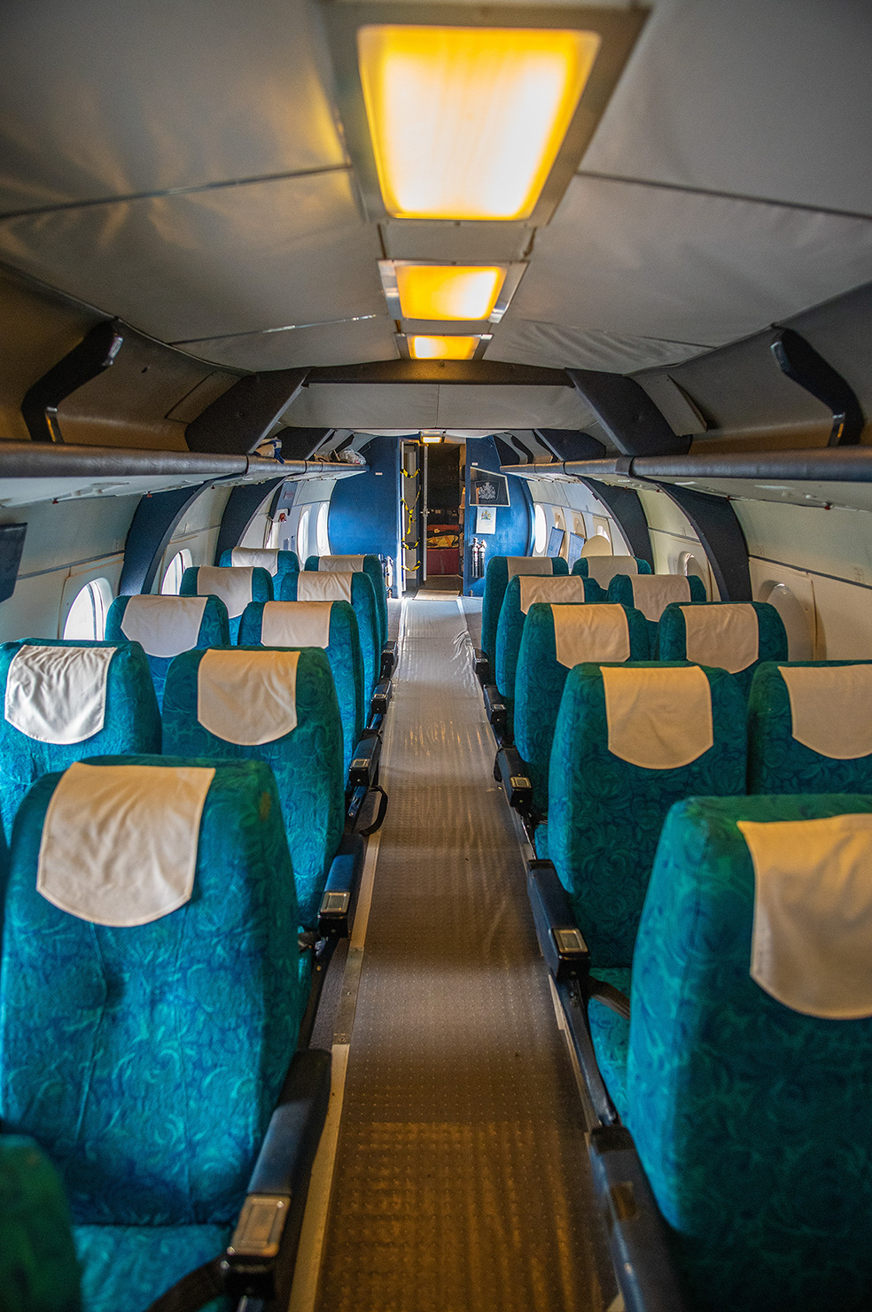 The image shows the interior of an airplane with rows of seats covered in blue and beige upholstery. The cabin is well-lit with overhead lights, and the aisle is clear, leading towards the front of the plane. The seats appear to be arranged in a 2-2 configuration, and the overall condition of the cabin looks clean and well-maintained.