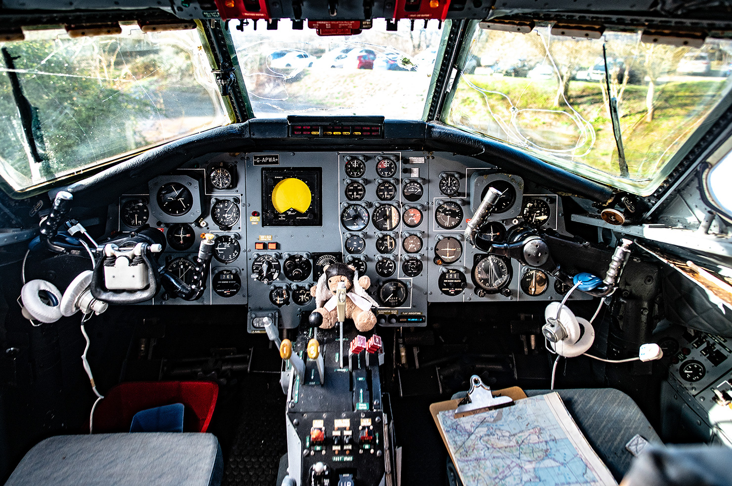 The image shows the cockpit of an aircraft, featuring a complex array of instruments and controls. The view through the windshield reveals a parking area with several cars and a natural landscape. Notably, there is a teddy bear placed in the center console, adding a touch of whimsy to the otherwise technical environment. The cockpit appears to be well-used, with various tools and a map visible, indicating it is an active or recently used aircraft. The overall scene suggests a blend of advanced aviation technology and personal touches.