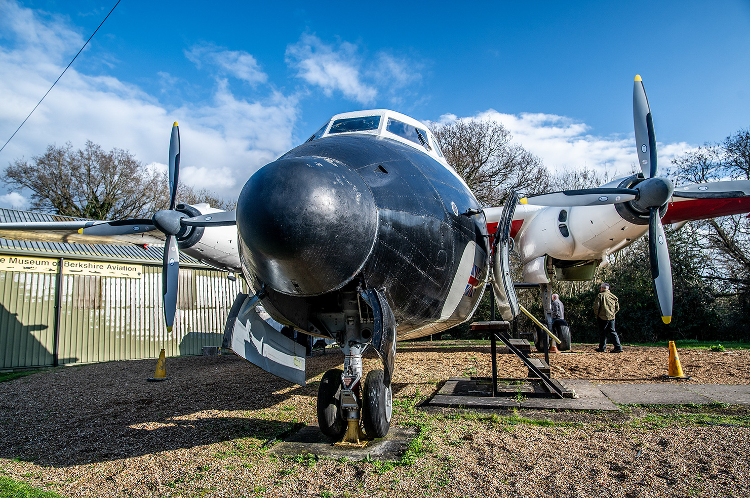 The image shows a vintage aircraft on display at the Museum of Berkshire Aviation. The aircraft is a twin-engine propeller plane with a distinctive black nose and silver body. It is positioned on a grassy area with its landing gear visible, and there are two people walking nearby. The museum building is in the background, and the sky is clear with some clouds.