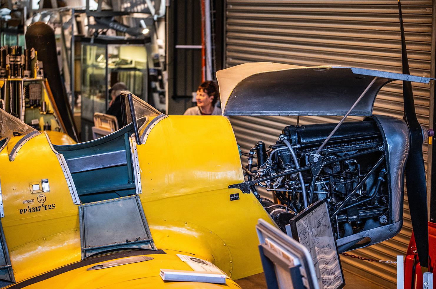 The image shows a vintage yellow biplane with its engine exposed, likely on display in a museum or exhibition setting. The aircraft's registration number PP4317 T2S is visible on the side. The engine is detailed with various components visible, and there are informational placards placed around the plane, suggesting it is part of an exhibit. A person is seen in the background, possibly a visitor or staff member, and the setting appears to be indoors with a closed shutter in the background.