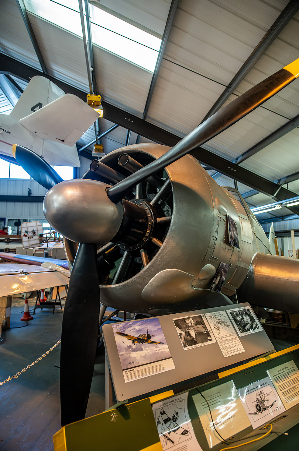 The image depicts an aircraft displayed in a museum setting. The aircraft is suspended from the ceiling, showcasing its propeller and front section. Below the aircraft, there are several informational panels with text and images, providing historical and technical details about the aircraft.