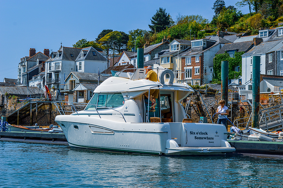 The image shows a picturesque harbor scene with a white yacht named 'Six o'clock Somewhere' docked at a marina. The yacht is moored next to a wooden pier, with a person standing nearby. In the background, there are charming houses and buildings on a hillside, surrounded by lush greenery. The weather appears to be clear and sunny, creating a serene and inviting atmosphere.