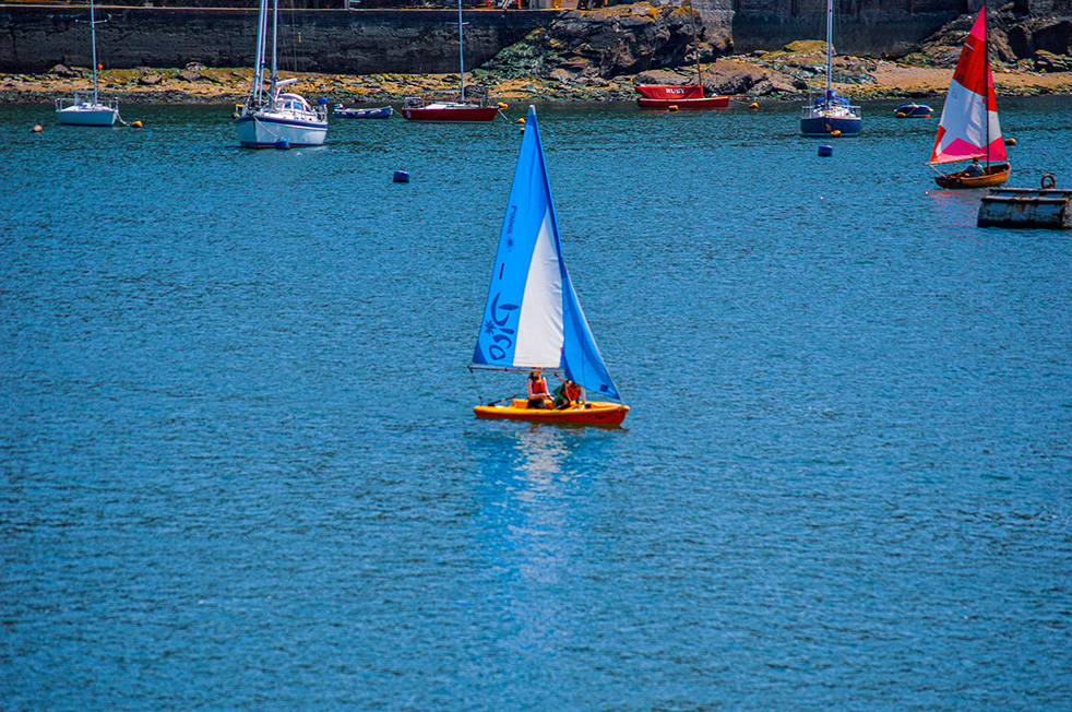 The image shows a small sailboat with a blue sail and the number 180 on it, sailing in a harbor. The boat has a bright orange hull and is carrying two people. In the background, there are several other boats, including sailboats and small motorboats, anchored near the shore. The water is a clear blue, and the scene appears to be set in a calm, sunny environment.