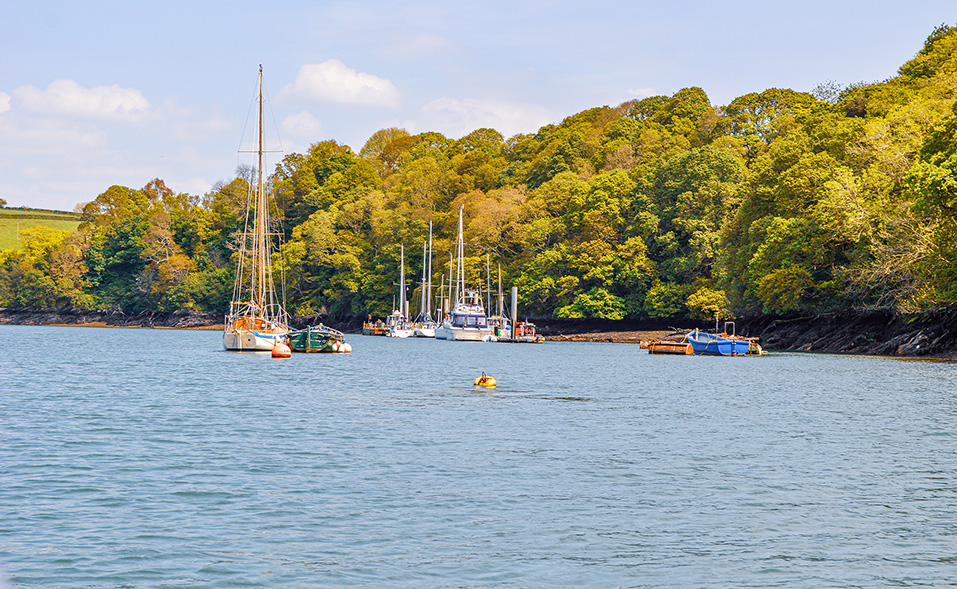 The image depicts a serene waterfront scene with several boats anchored near a lush, green forested area. The water is calm, and the sky is clear with a few clouds. The boats vary in size and type, including sailboats and smaller vessels. The shoreline is rocky, and the trees appear to be in full foliage, suggesting it might be spring or summer. The overall atmosphere is peaceful and picturesque.