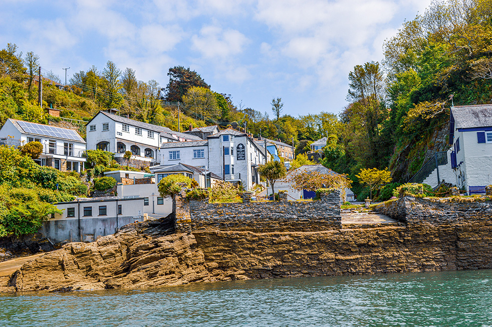 The image depicts a picturesque coastal village with houses built on a hillside, overlooking a rocky shoreline with calm blue-green waters. The houses are nestled among lush greenery and trees, creating a serene and scenic environment. The architecture varies, with some modern and some traditional styles, blending harmoniously with the natural landscape. The sky is clear with some scattered clouds, adding to the overall tranquility of the scene.