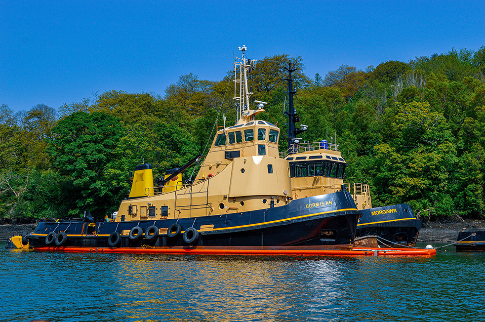 The image depicts a large, yellow and black tugboat named 'Morgawr' docked near a wooded area. The boat is equipped with various maritime equipment and is situated in calm waters with a red bumper along its side.