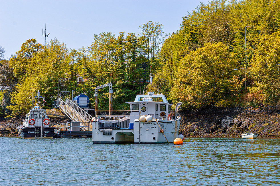 The image depicts a serene waterfront scene with two boats docked near a small pier. The larger boat, named 'GREEN ISLE,' is positioned centrally, while a smaller boat, 'FOWEY,' is to the left. The area is surrounded by lush greenery and trees, with a rocky shore in the background. The sky is clear, suggesting a calm, sunny day. The overall atmosphere is peaceful and picturesque.