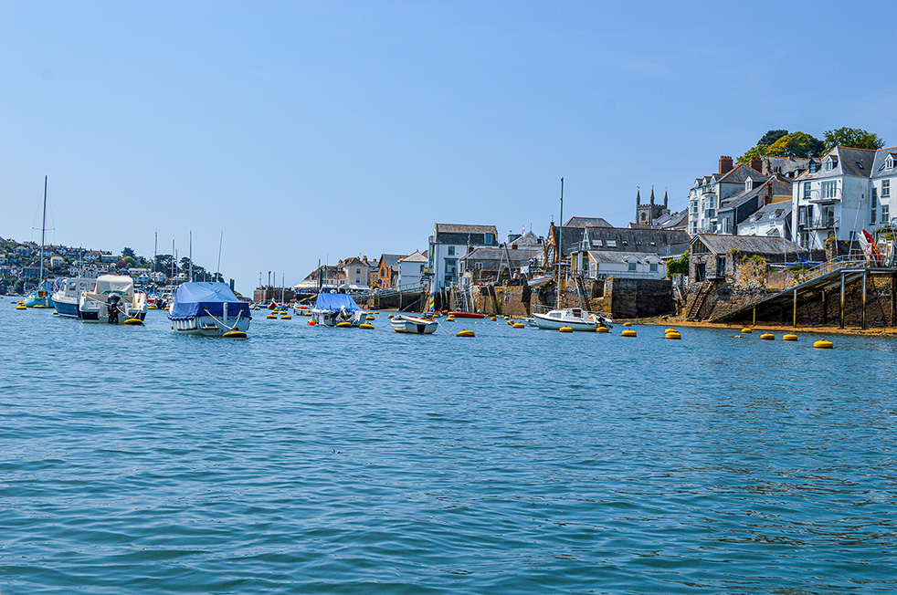 This image captures a vibrant coastal scene, showcasing a hillside town densely populated with a variety of architectural styles. Buildings range from grand, multi-story hotels and residences with ornate detailing to charming, smaller homes, all nestled amidst lush greenery. The town overlooks a serene, blue body of water, with a rocky shoreline in the foreground featuring a small beach and a red buoy. The clear blue sky suggests a pleasant, sunny day, creating a tranquil and inviting atmosphere